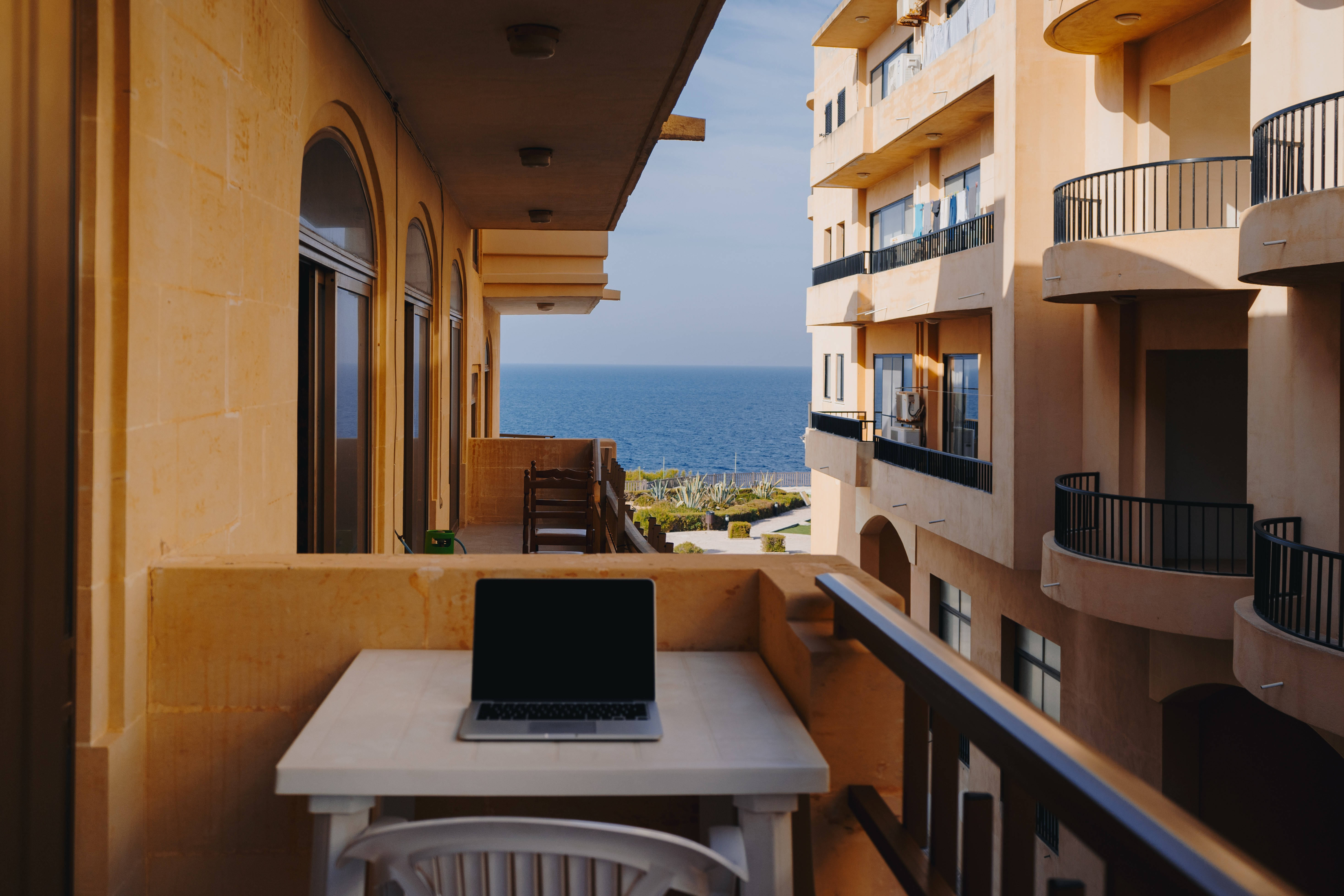 Malta 桌布 壁紙 — 一台筆記型電腦放在白色桌子上，位於陽光普照的陽台，俯瞰藍色海洋，附近有米色公寓大樓。