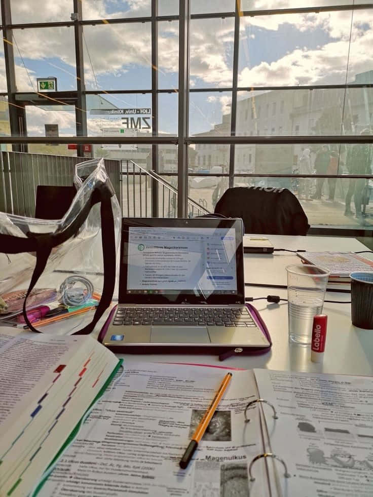 書桌 桌布 壁紙 — 一台筆記型電腦放在雜亂的書桌上，有打開的筆記本和一杯水，透過大窗戶看到明亮的城市景觀。