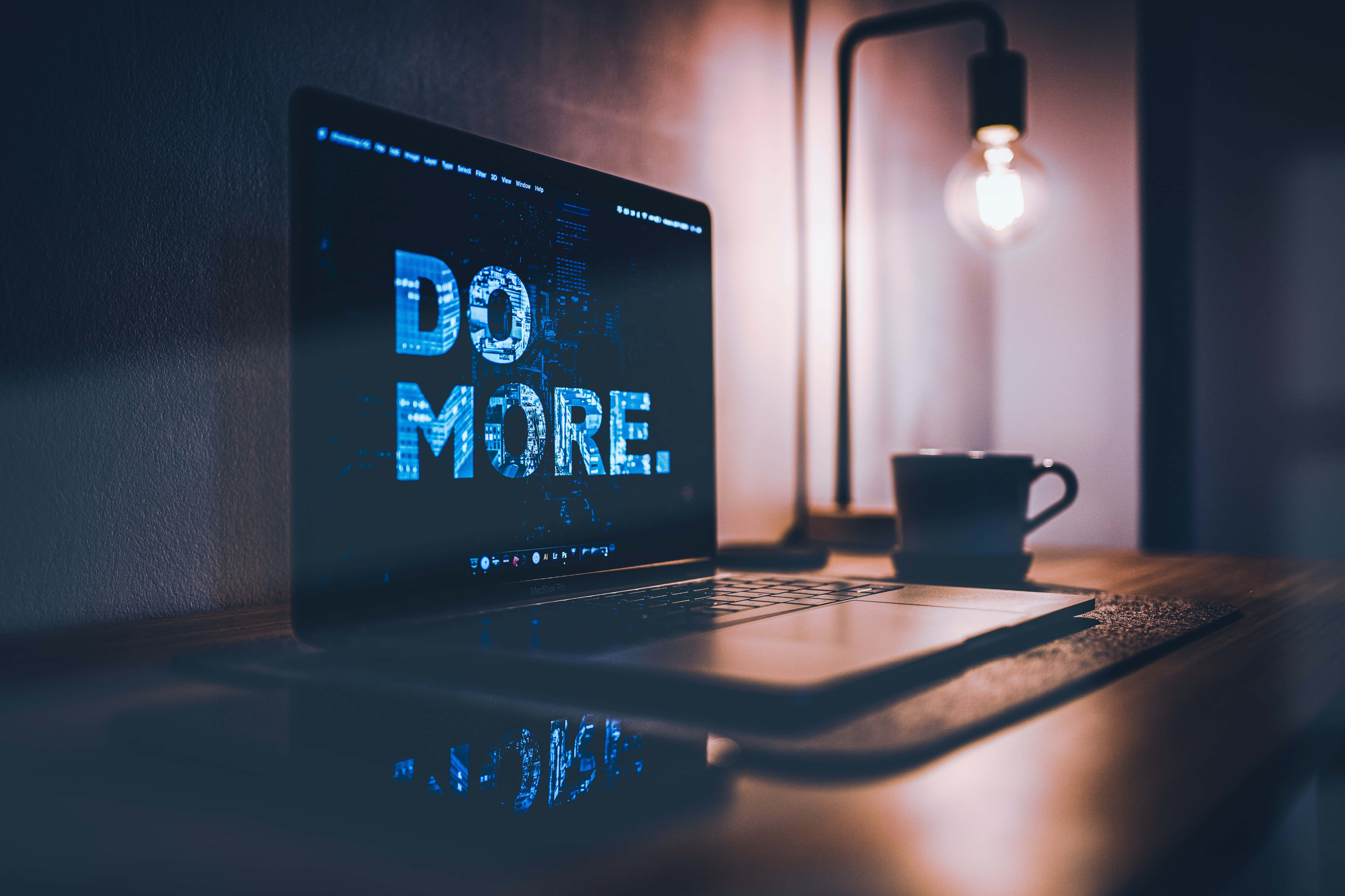 筆電 桌布 壁紙 — 一台筆記型電腦顯示發光藍色文字「DO MORE.」，放在木桌上，旁邊有一盞亮著的燈和咖啡杯，房間燈光昏暗。