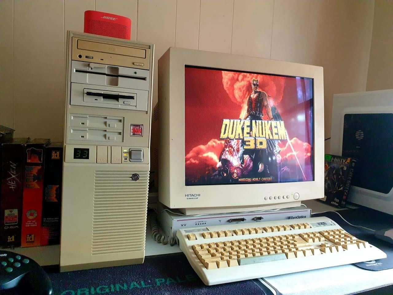 Old Pc 桌布 壁紙 — 一台米色復古桌上型電腦在其CRT顯示器上顯示Duke Nukem 3D，周圍環繞著遊戲配件和CD-ROM盒。
