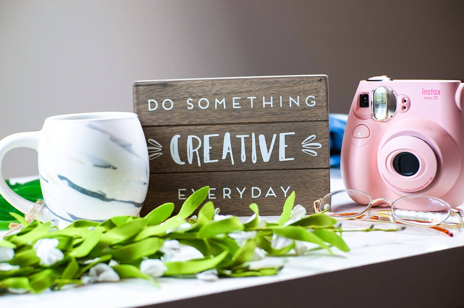 Motivational Quotes 桌布 壁紙 — 一台粉紅色拍立得相機和一個大理石馬克杯放在一個木牌旁邊，木牌上寫著「DO SOMETHING CREATIVE EVERYDAY」，周圍環繞著新鮮的綠色植物。