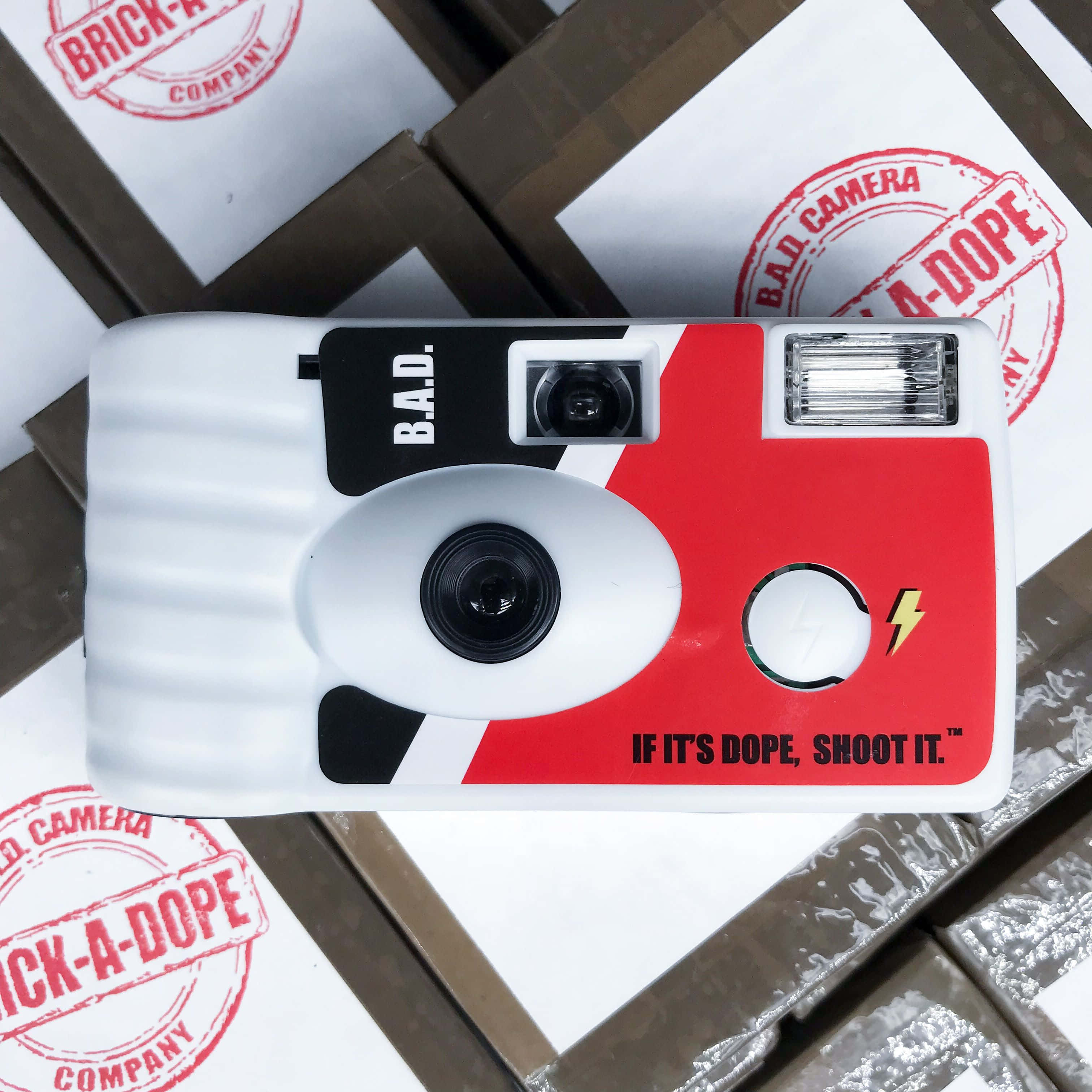 Disposable Camera 桌布 壁紙 — 一台紅白相間、印有 B.A.D. 標誌的即時相機，放在印有 'BRICK-A-DOPE' 印章的品牌紙箱上。