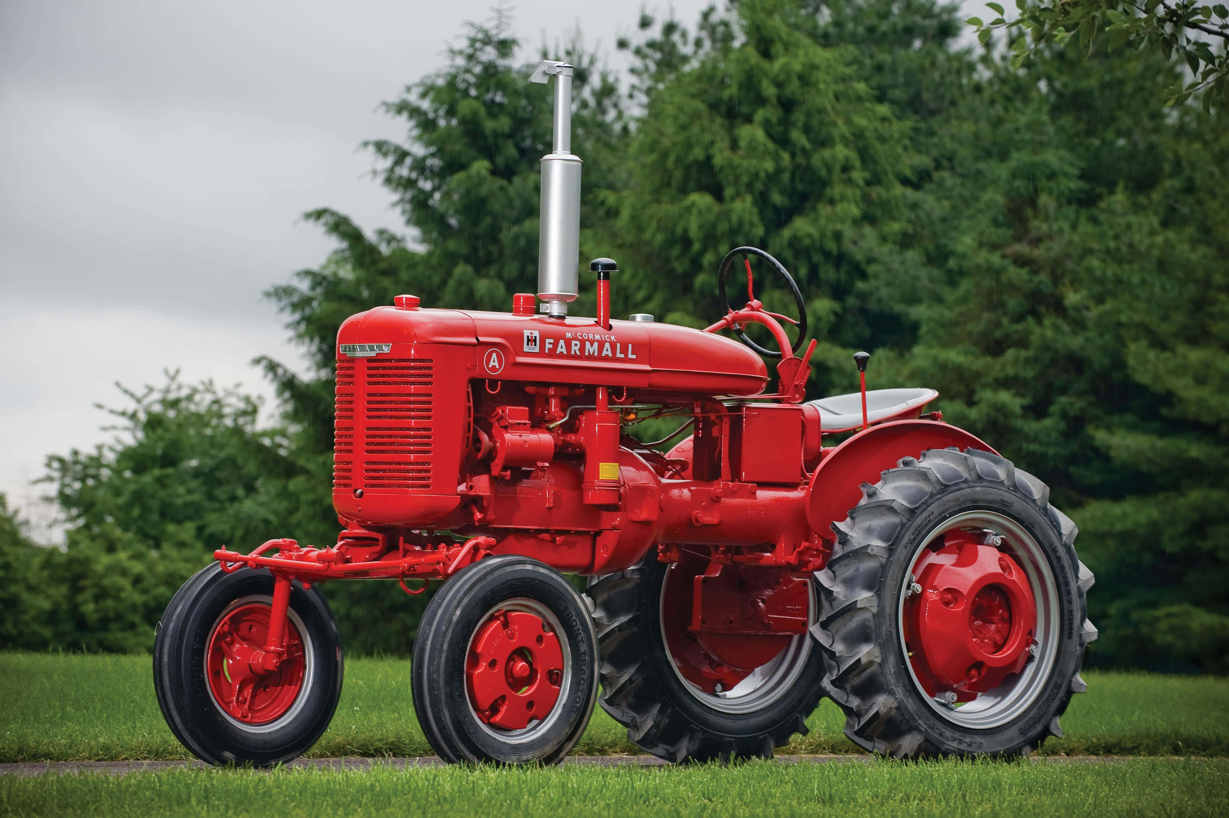 Tractor 桌布 壁紙 — 一台經典紅色 Farmall 拖拉機自豪地矗立在草地上，背後是茂密的綠樹，天空多雲。