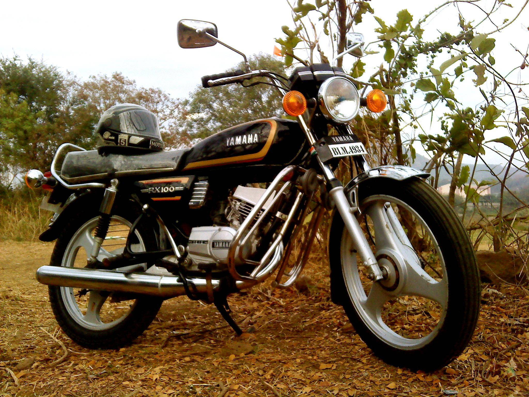 Rx100 桌布 壁紙 — 一台經典黑色Yamaha RX100機車停在乾葉上，旁邊有綠色灌木，天空多雲。