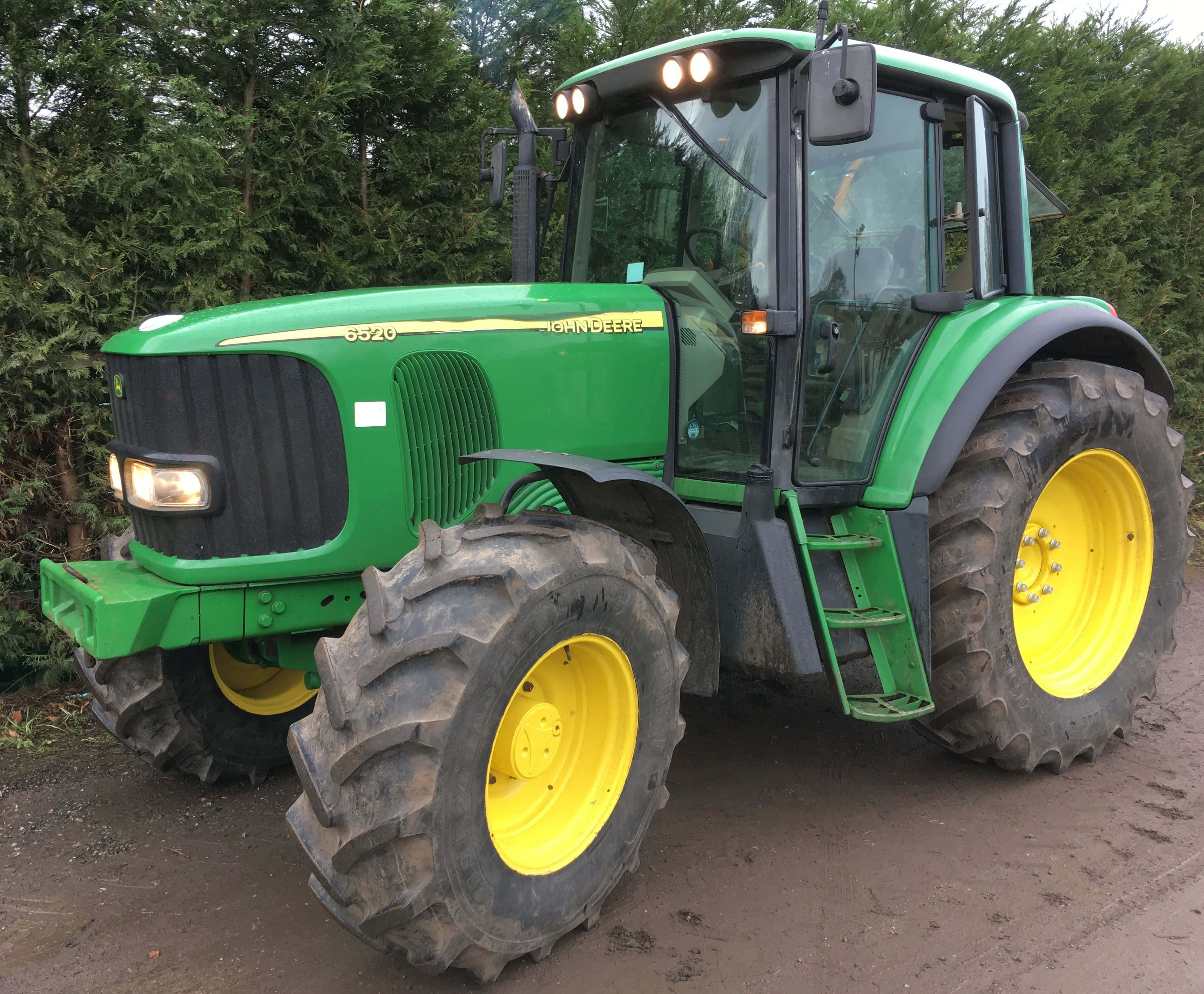 Tractor 桌布 壁紙 — 一台綠色和黃色的John Deere 6520拖拉機，帶有大型泥濘輪胎，停放在常綠樹附近的泥土上。