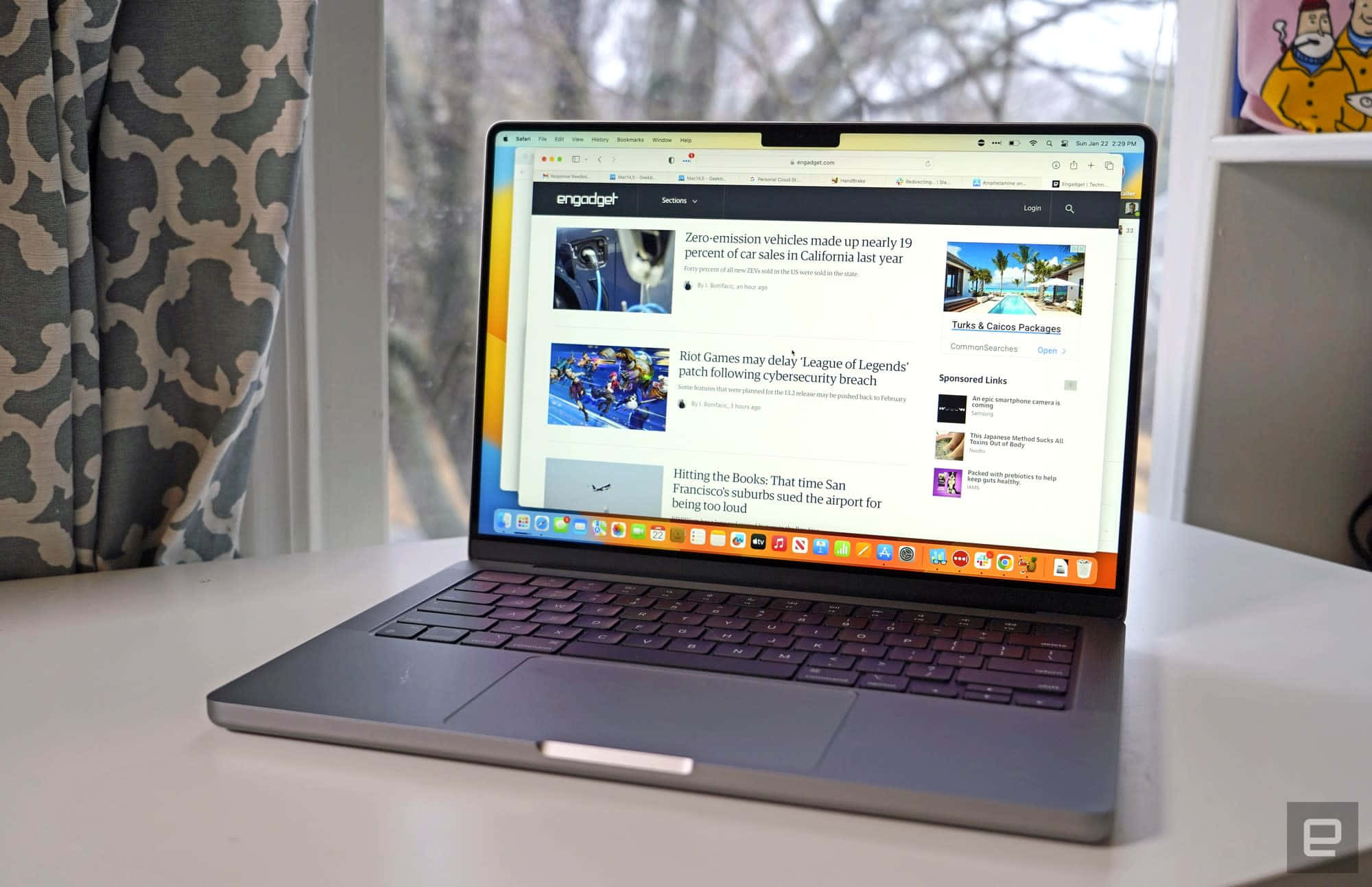 MacBook 動態圖片 桌布 壁紙 — 一台纖薄的銀色筆記型電腦打開放在白色桌子上，顯示Engadget新聞頁面，有關於汽車和遊戲的文章，旁邊有圖案窗簾和卡通海報。