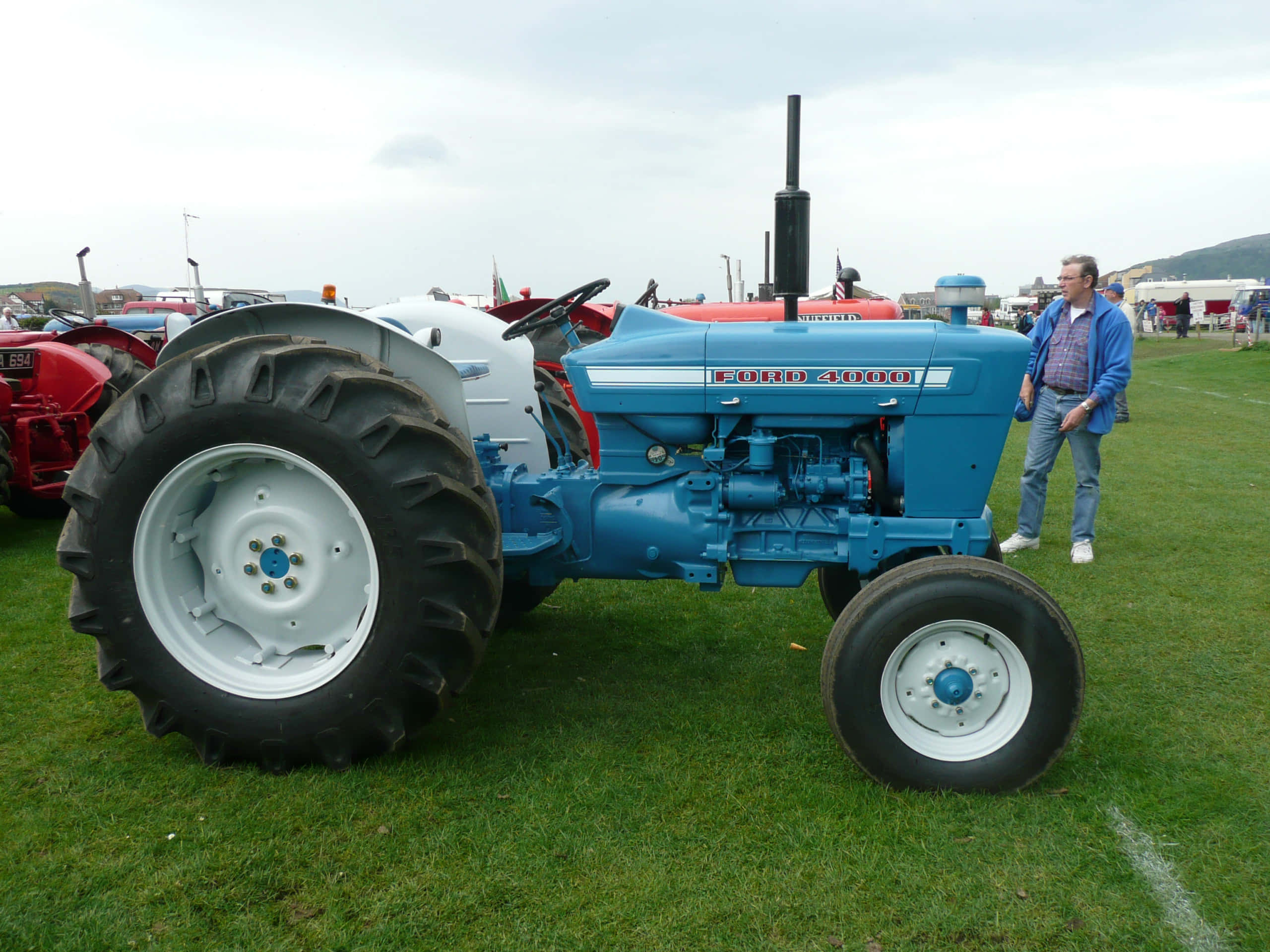 Tractor 桌布 壁紙 — 一台藍色復古福特4000拖拉機，白色車輪，停在綠色草地上，旁邊有一個男人，在戶外農業展覽上。