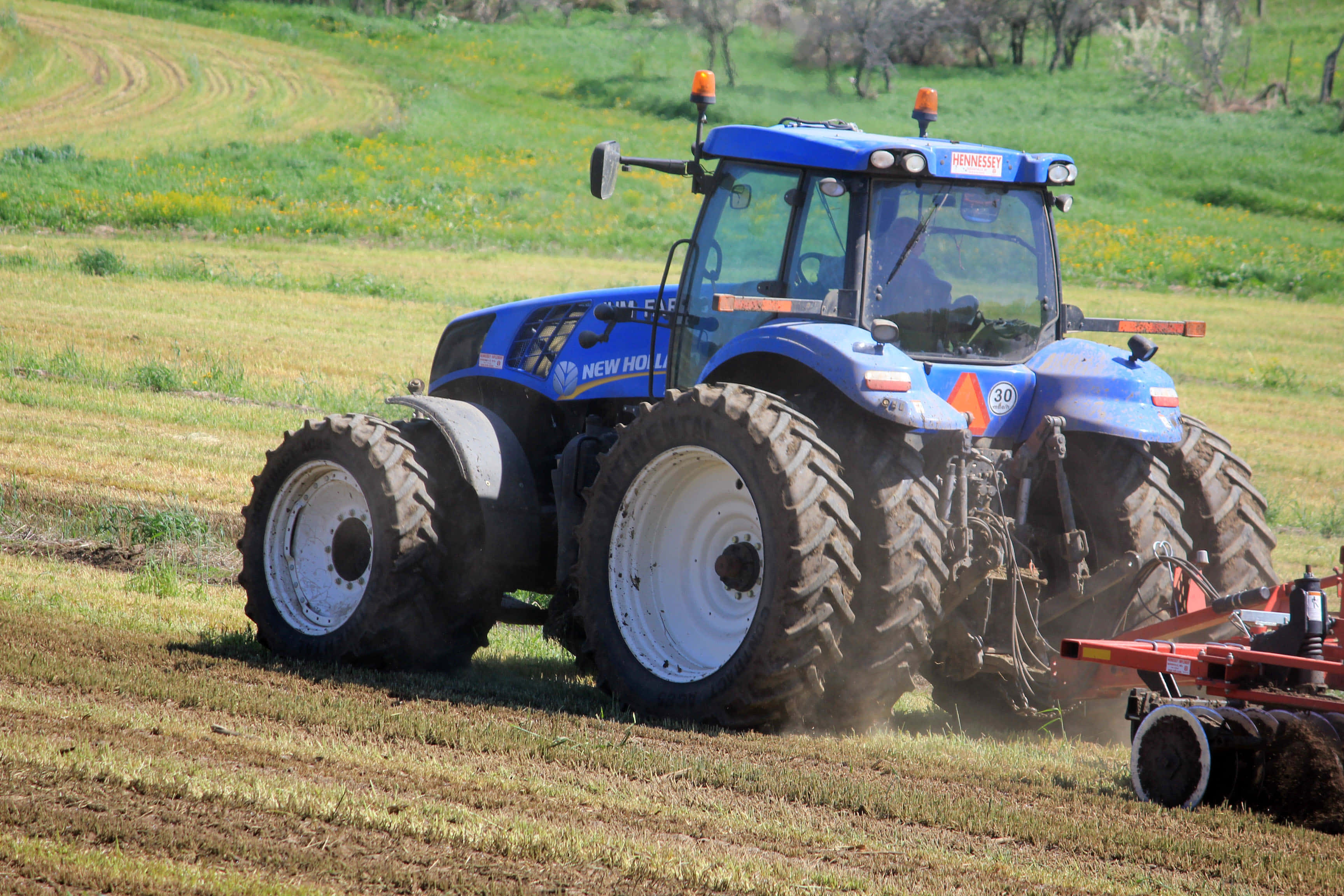 Tractor 桌布 壁紙 — 一台藍色紐荷蘭拖拉機在陽光明媚的綠色田野中犁地，留下整齊的犁溝。