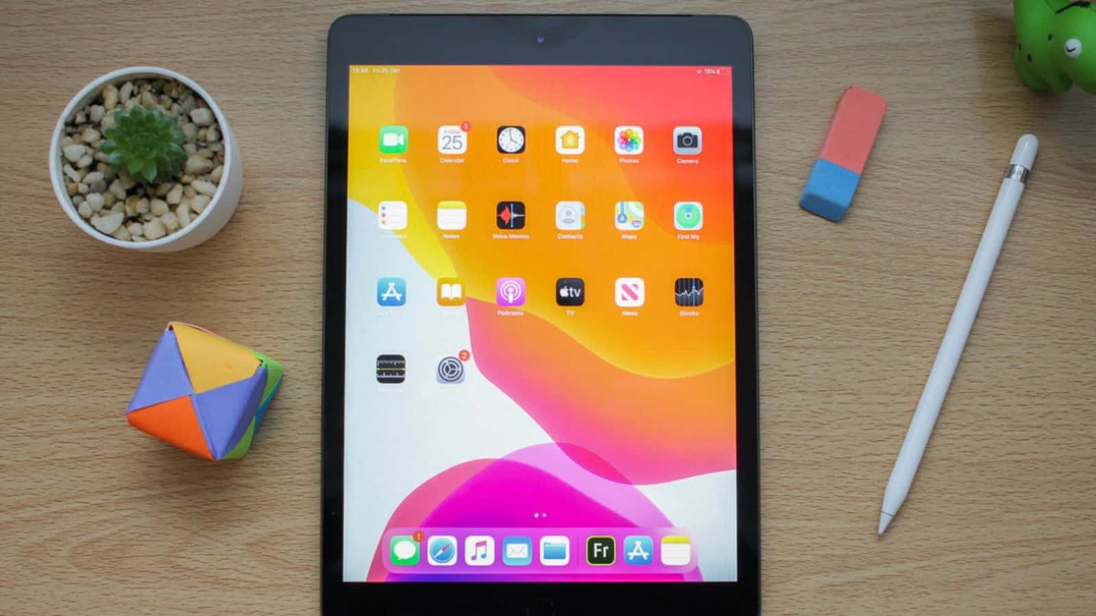 iPad 桌布 壁紙 — 一台裝有彩色應用程式的平板電腦放在木桌上，旁邊有盆栽、摺紙、鉛筆和橡皮擦。