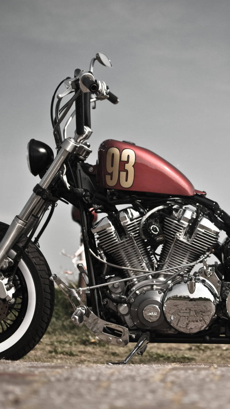Bobber 桌布 壁紙 — 一台訂製紅色機車，帶有鍍鉻細節，油箱上印有93號，停在碎石地面上，天空多雲。