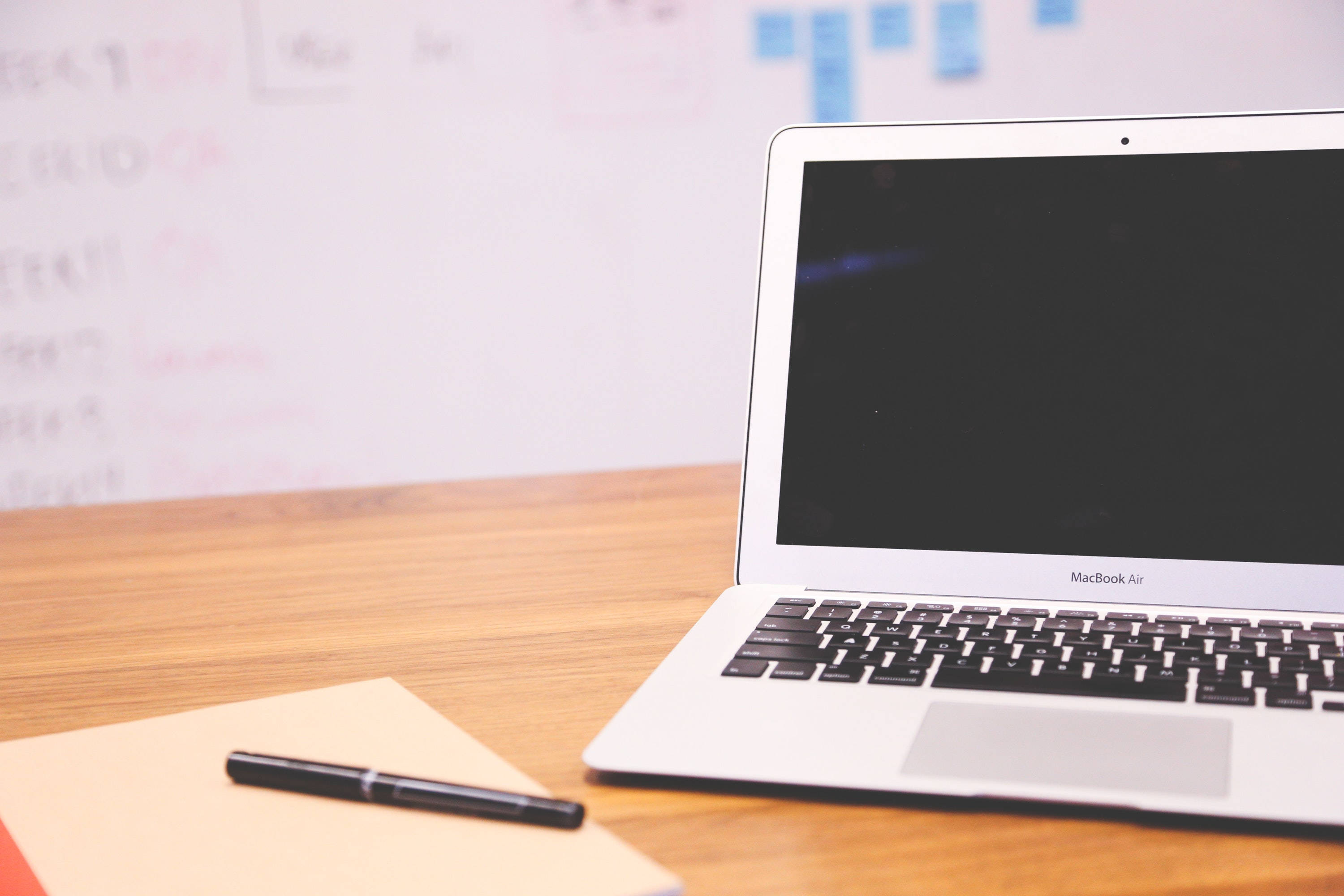 Cute Laptop 桌布 壁紙 — 一台銀色 MacBook Air 打開放在木桌上，旁邊有筆記本和筆，背景有模糊的白板。