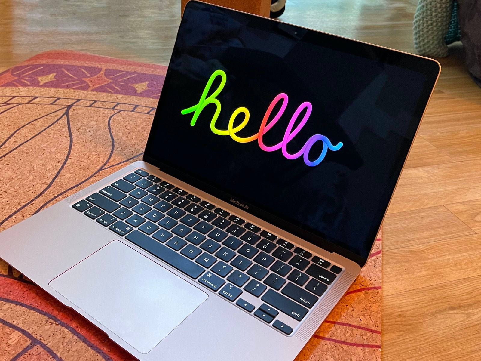 Hello 桌布 壁紙 — 一台銀色 MacBook Air 在木地板上的圖案地毯上，螢幕顯示彩虹漸變的哈囉。