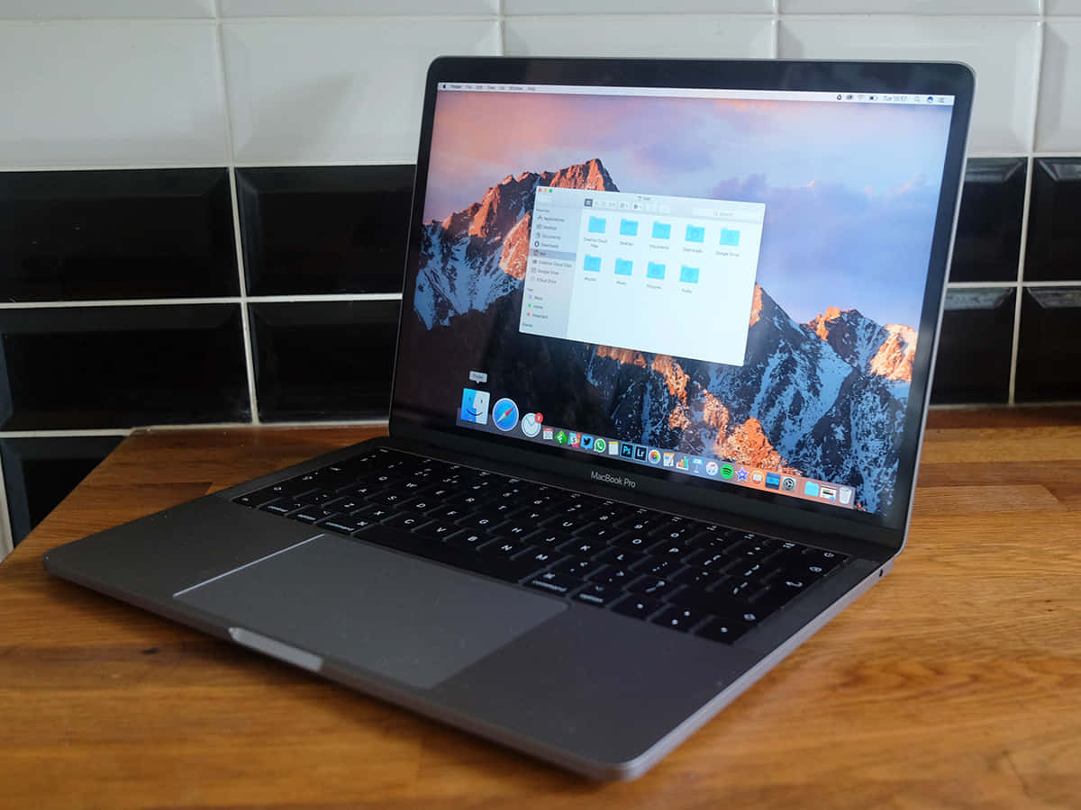 MacBook 動態圖片 桌布 壁紙 — 一台銀色 MacBook Pro 打開放在木桌上，背景是黑色瓷磚牆，螢幕上顯示山脈桌布。