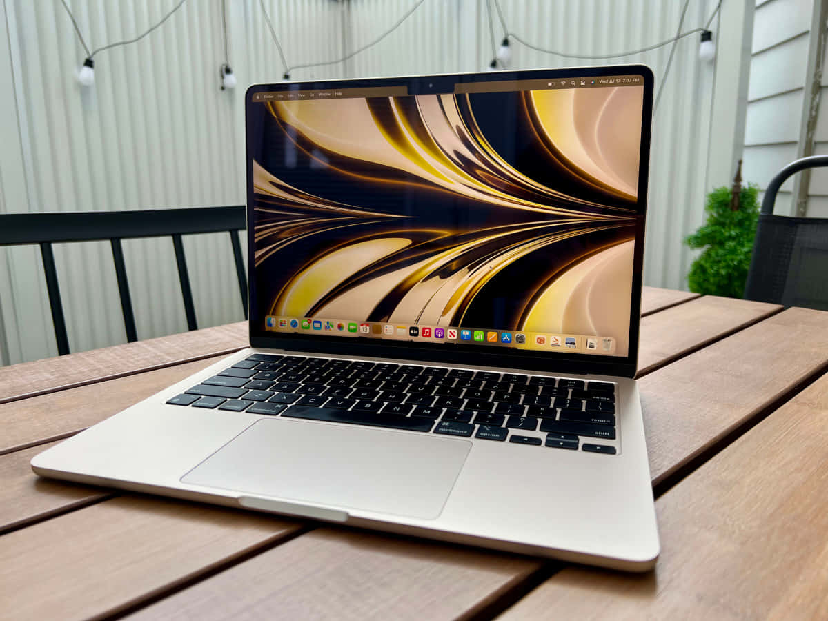 MacBook 動態圖片 桌布 壁紙 — 一台銀色筆記型電腦，顯示著旋轉的金色和黑色桌布，打開放在木製露台桌上，上方有燈串。