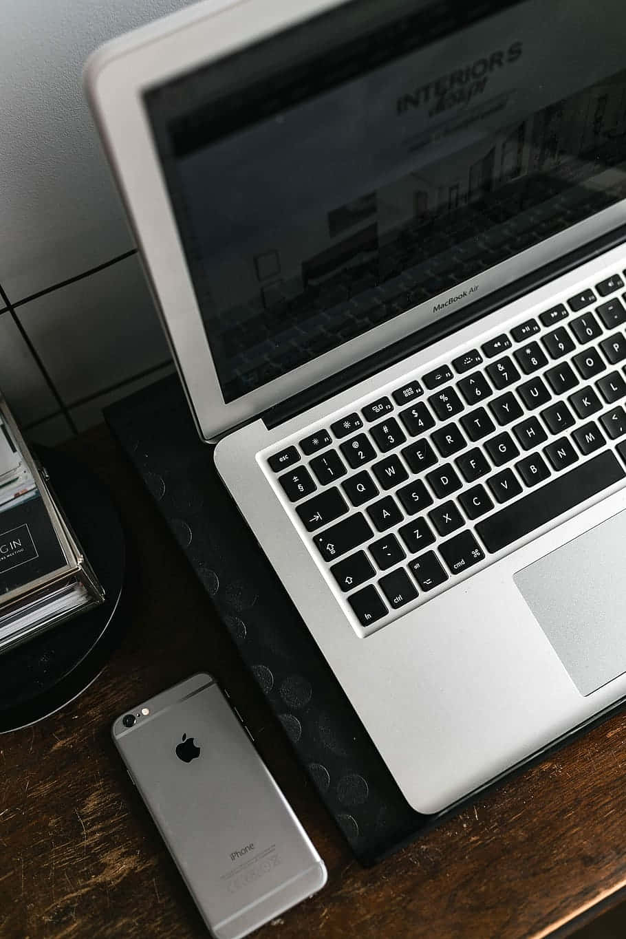 Desk 桌布 壁紙 — 一台銀色筆記型電腦和iPhone放在深色木桌上，旁邊有一塊黑色紋理墊和一疊雜誌。