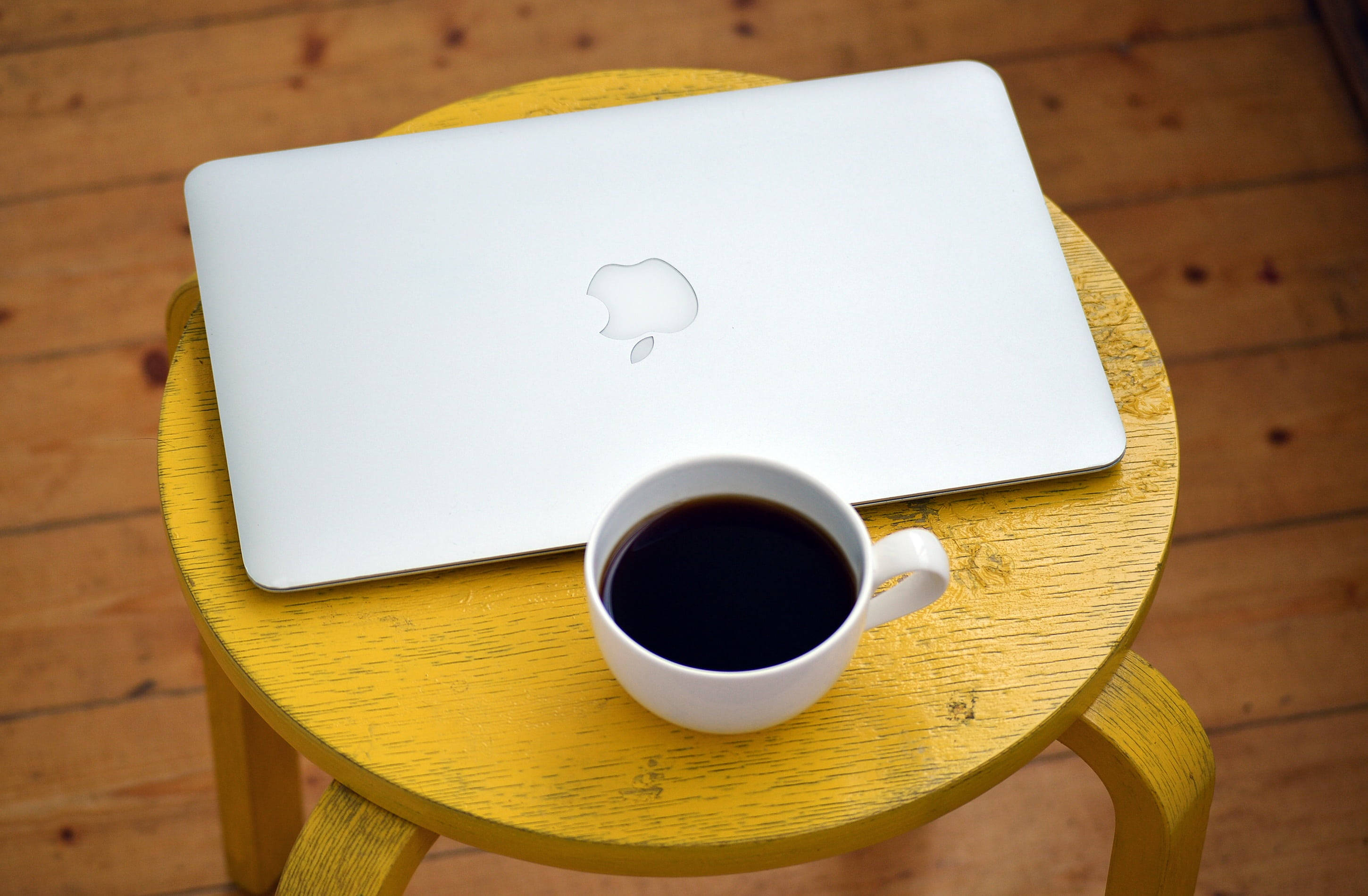 Coffee Aesthetic 桌布 壁紙 — 一台銀色筆記型電腦和一個白色咖啡杯放在一個亮黃色木凳上，背景是溫暖的木地板。