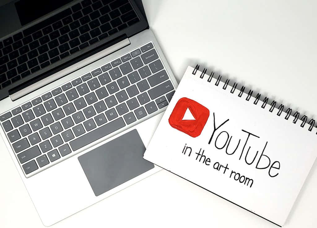 YouTube 桌布 壁紙 — 一台銀色筆記型電腦和一本帶有手繪YouTube標誌的螺旋筆記本，放在潔白的表面上。