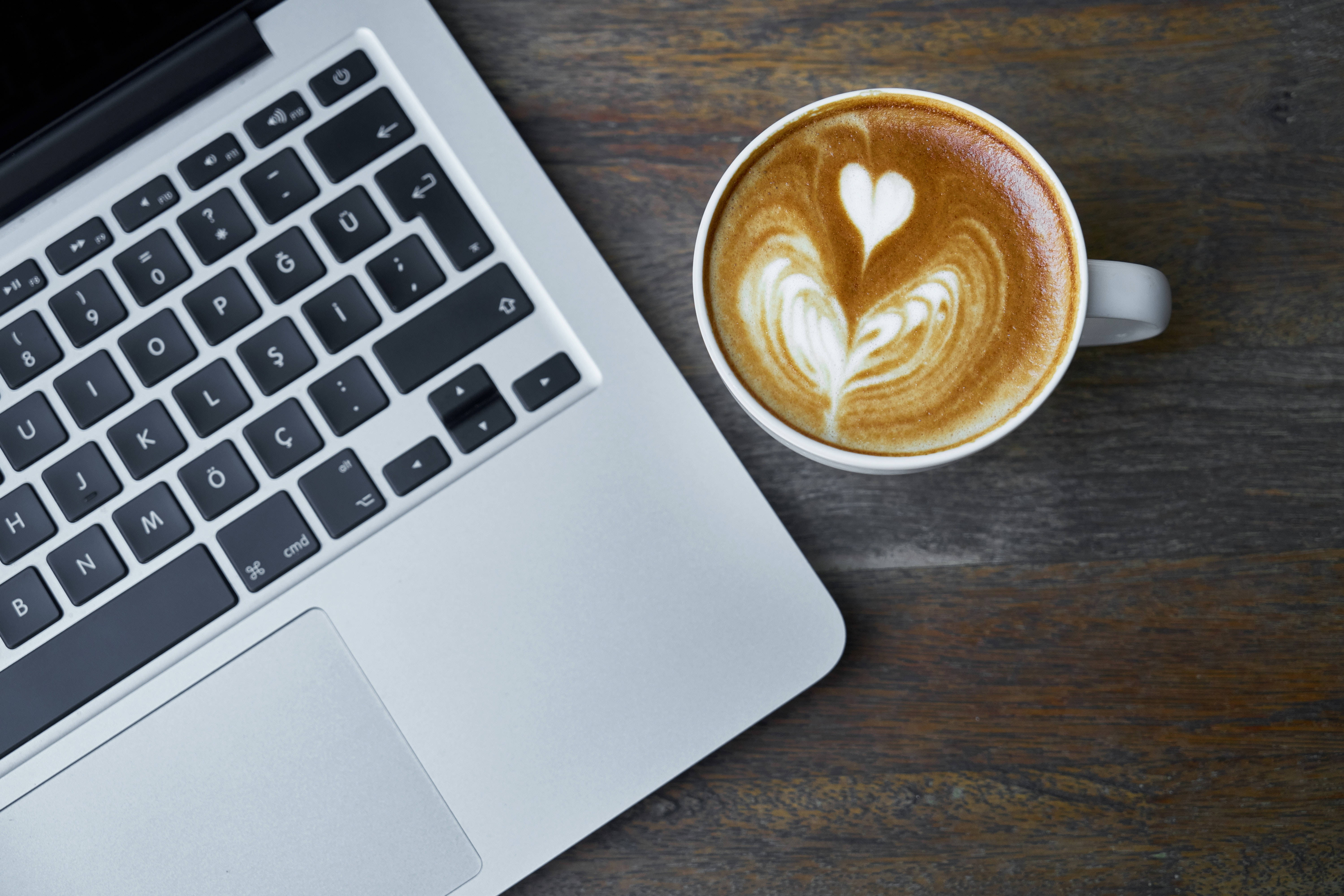 MacBook 動態圖片 桌布 壁紙 — 一台銀色筆記型電腦和一杯心形拉花咖啡放在深色木桌上。