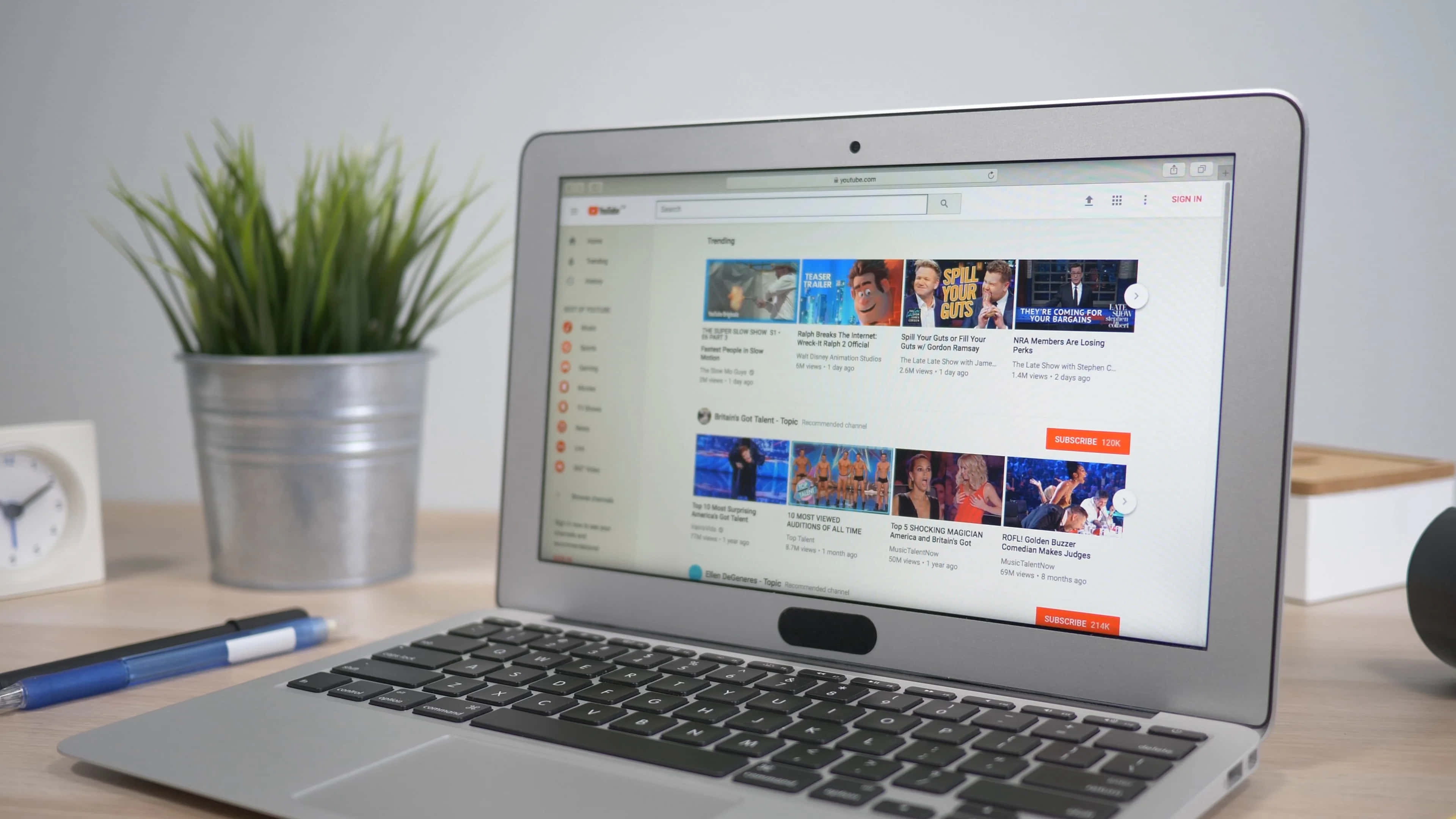 YouTube 桌布 壁紙 — 一台銀色筆記型電腦在木桌上顯示YouTube首頁，旁邊有盆栽植物和時鐘。
