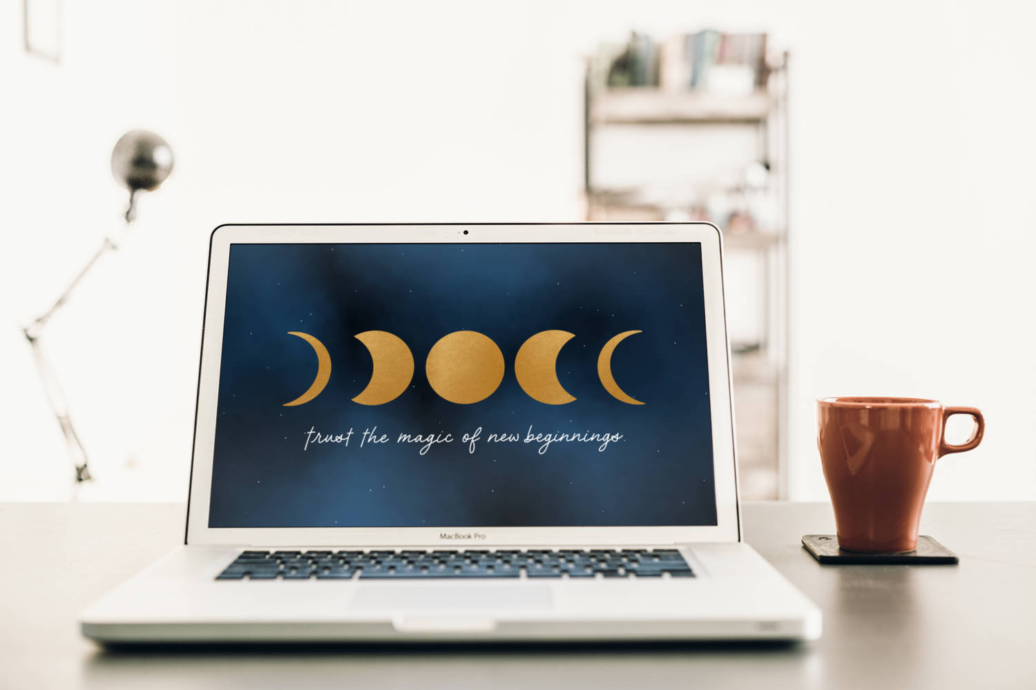 Working From Home 桌布 壁紙 — 一台銀色筆記型電腦在簡約的桌子上，旁邊有一個陶土杯，顯示金色的月相和鼓舞人心的文字。