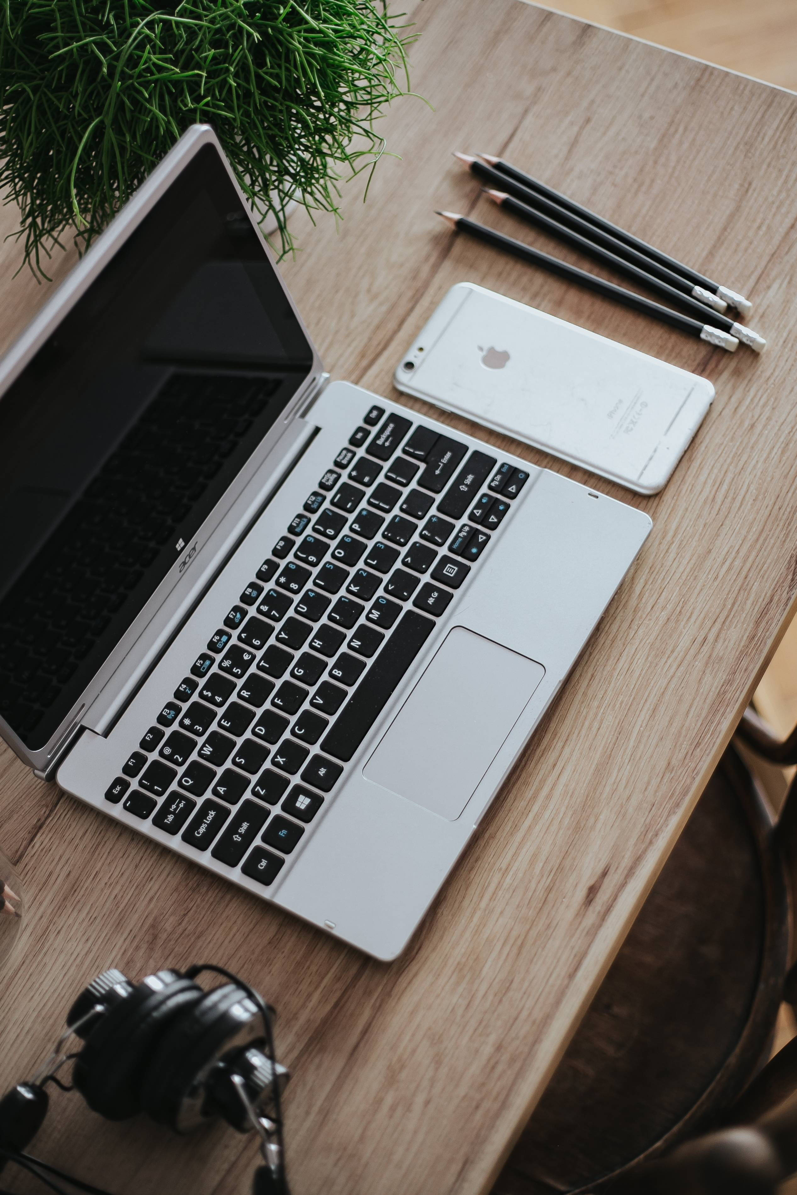 MacBook 動態圖片 桌布 壁紙 — 一台銀色筆記型電腦打開放在木桌上，旁邊有白色電話、鉛筆和耳機，附近有一棵綠色植物。