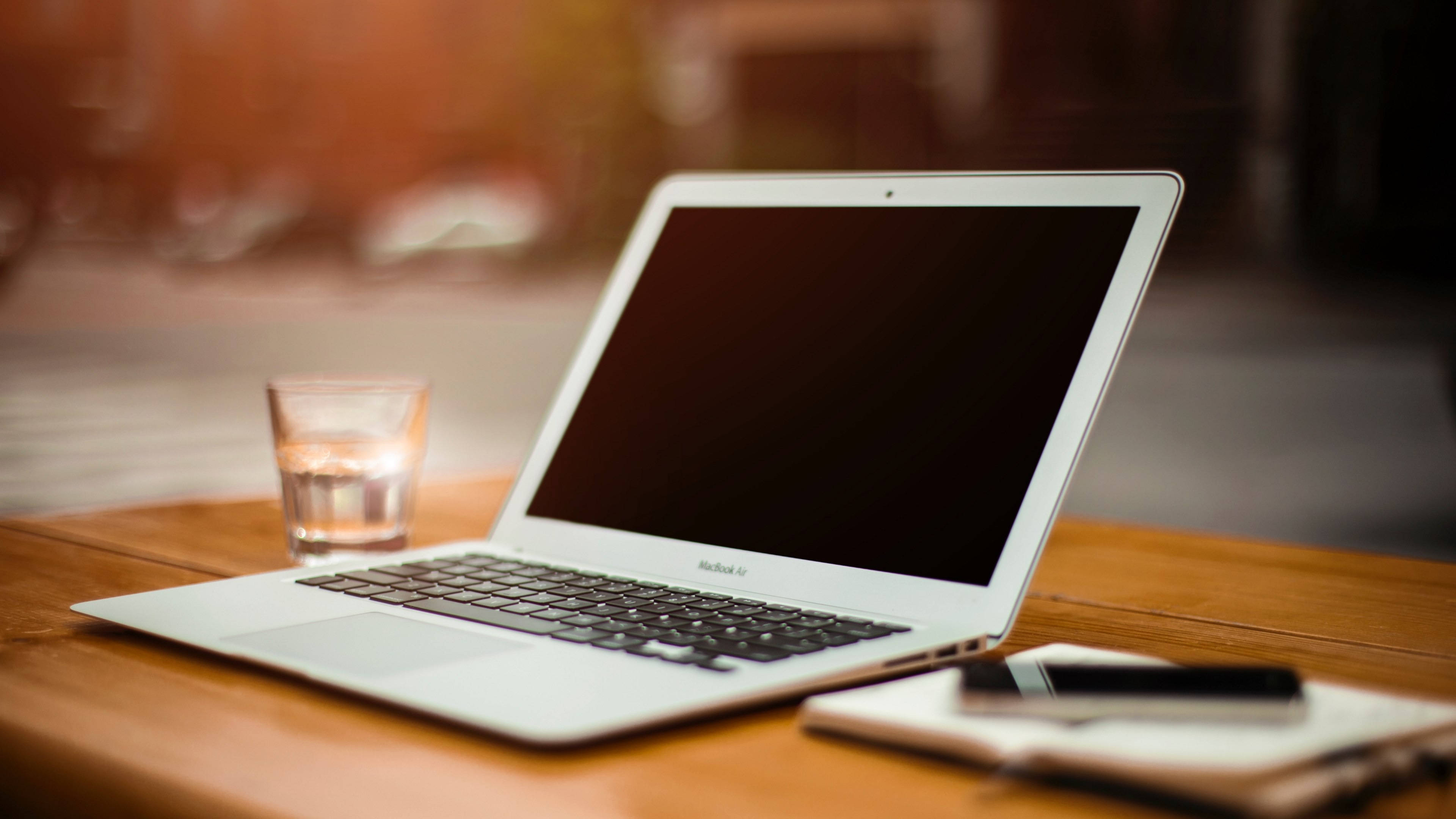 MacBook 動態圖片 桌布 壁紙 — 一台銀色筆記型電腦打開放在木桌上，旁邊有一杯水和一本帶筆的筆記本。