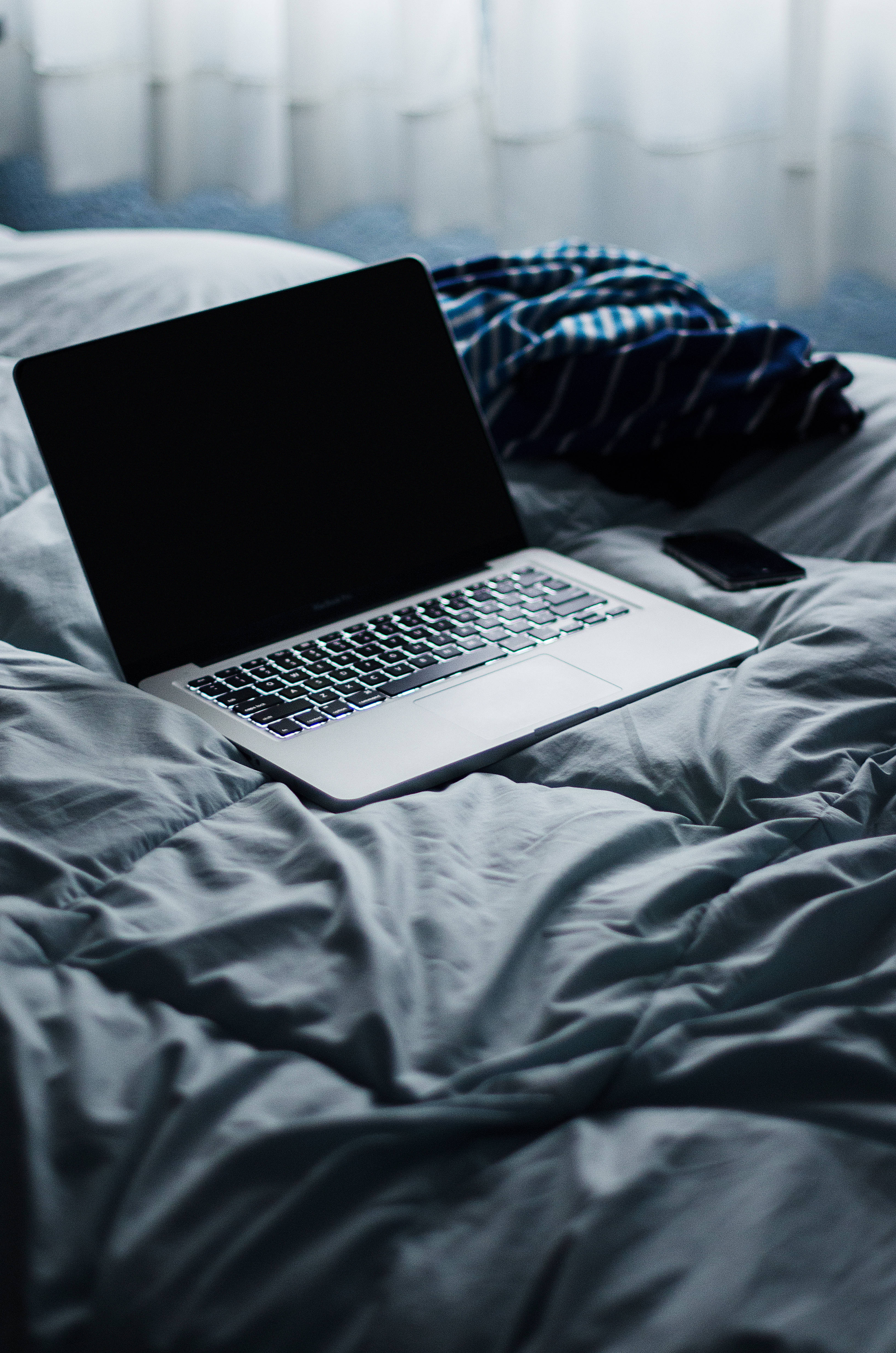 bed 桌布 壁紙 — 一台銀色筆記型電腦打開放在皺巴巴的灰色床單上，旁邊有一部智慧型手機和一件皺巴巴的藍色條紋襯衫，靠近有薄窗簾的窗戶。