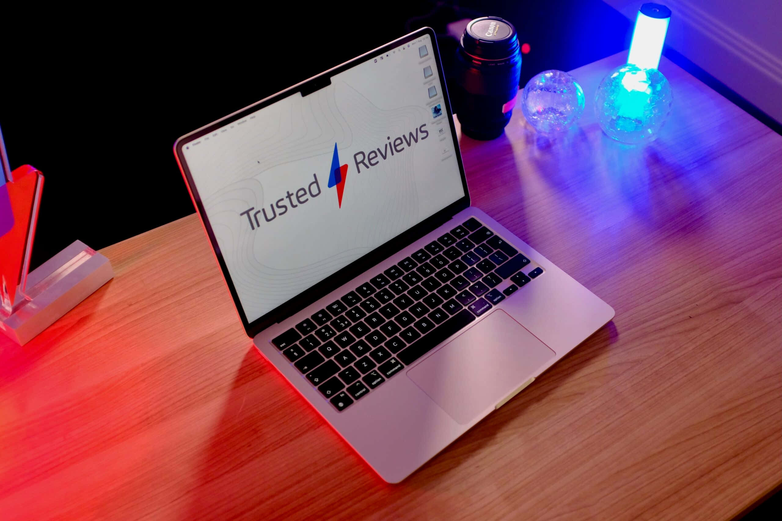 MacBook 動態圖片 桌布 壁紙 — 一台銀色筆記型電腦顯示 Trusted Reviews，放在木桌上，被紅色和藍色霓虹燈裝飾照亮。