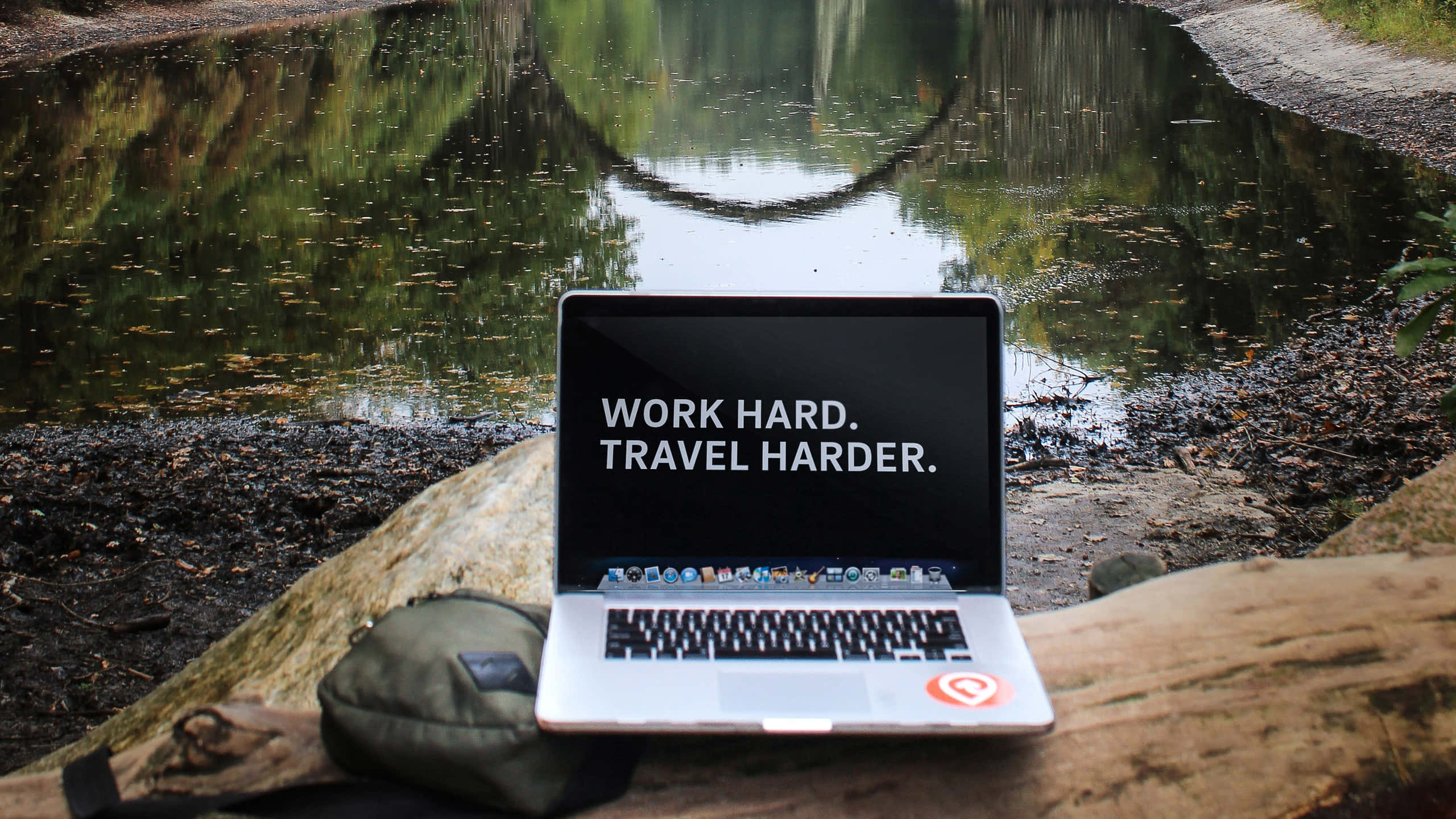 旅行 桌布 壁紙 — 一台銀色筆記型電腦顯示「WORK HARD. TRAVEL HARDER.」，放在一根圓木上，旁邊是倒映著樹木的寧靜森林池塘。
