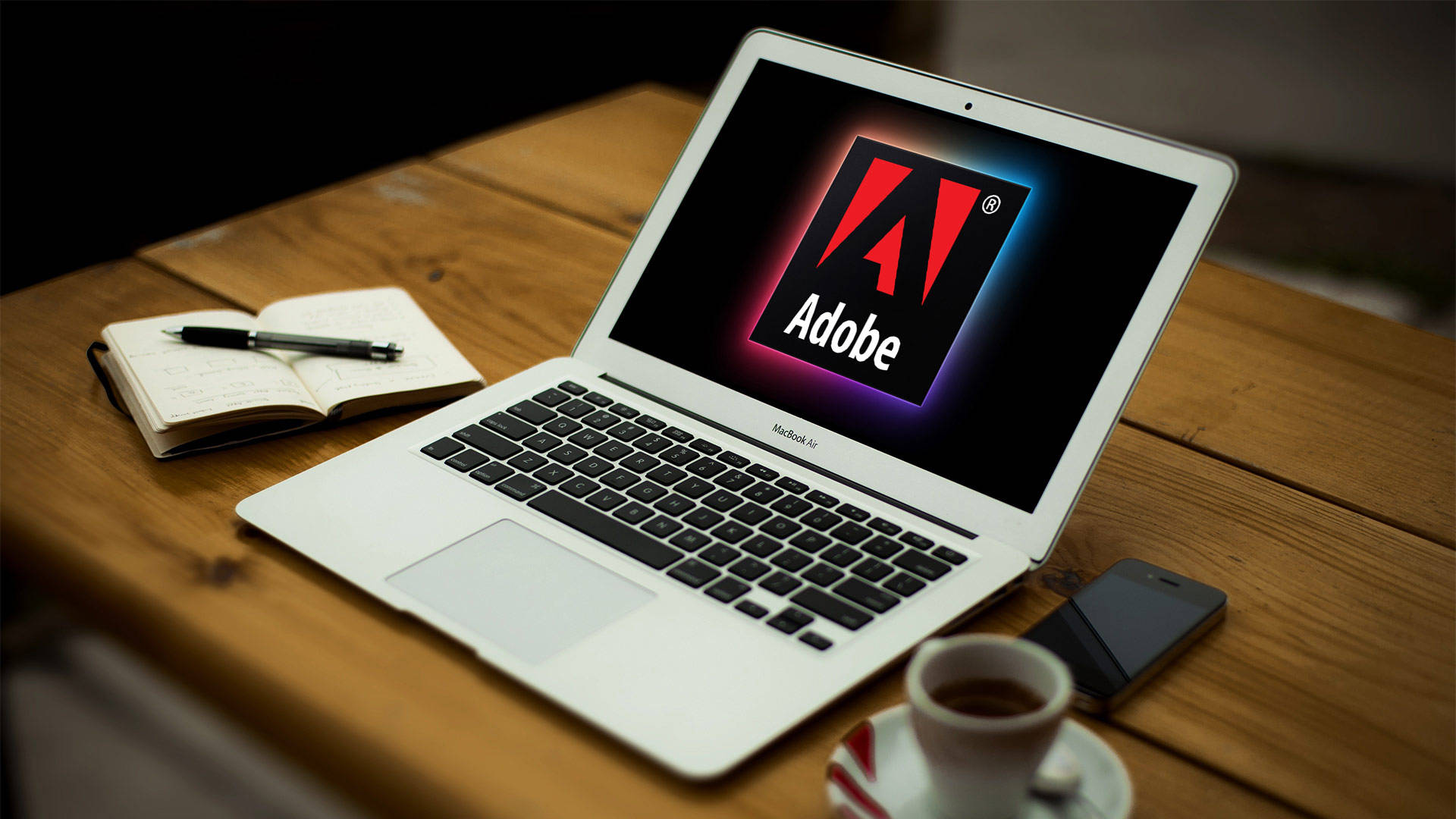 桌布 壁紙 — 一台銀色筆記型電腦顯示Adobe標誌，放在木桌上，旁邊有咖啡杯、筆記本和智慧型手機。