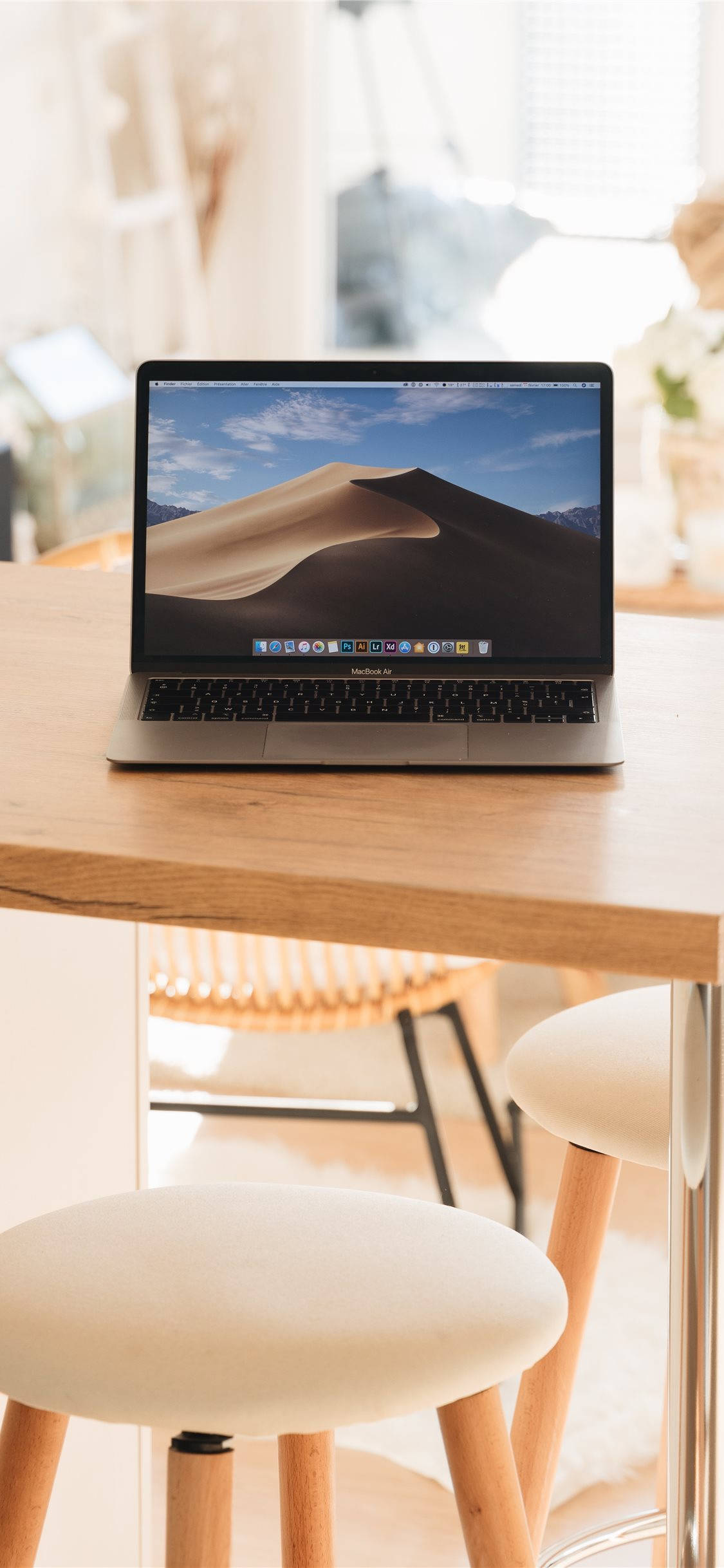 MacBook 動態圖片 桌布 壁紙 — 一台銀色筆記型電腦顯示沙漠沙丘桌布，放在淺色木桌上，搭配編織凳子，處於明亮的室內環境。