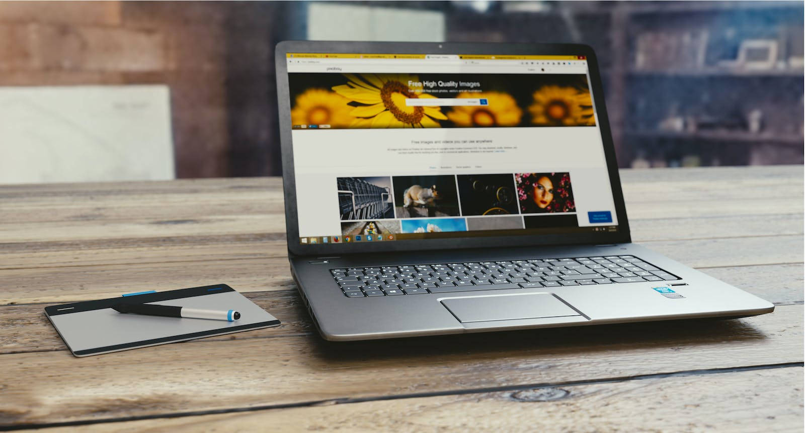 Website 桌布 壁紙 — 一台銀色筆記型電腦顯示著圖庫網站，放在一張質樸的木桌上，旁邊有一個繪圖板和觸控筆。