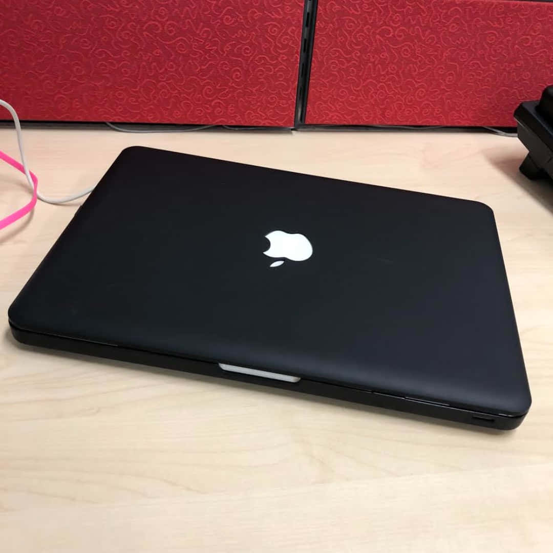 Black Macbook 桌布 壁紙 — 一台闔上的黑色MacBook放在淺色木桌上，後方有紅色圖案面板。