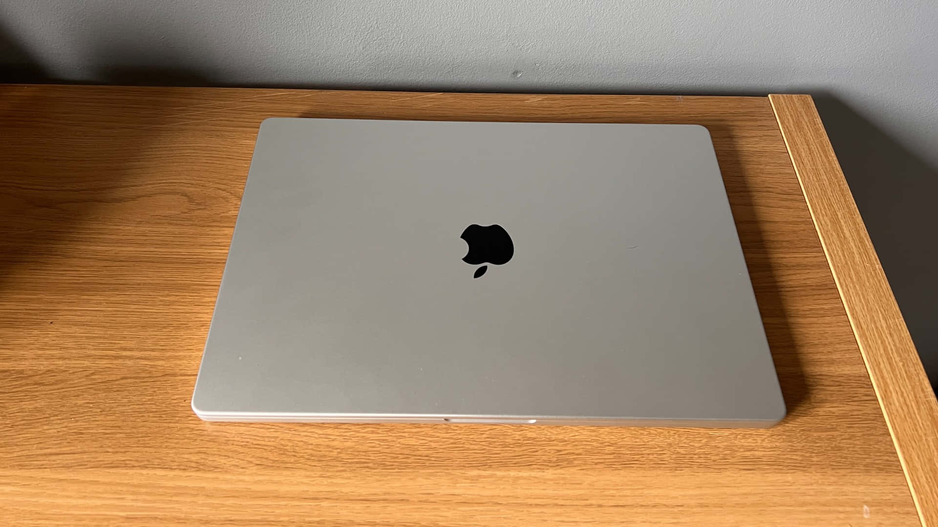 MacBook 動態圖片 桌布 壁紙 — 一台關閉的銀色MacBook放在淺色木桌上，背景是純灰色牆壁。