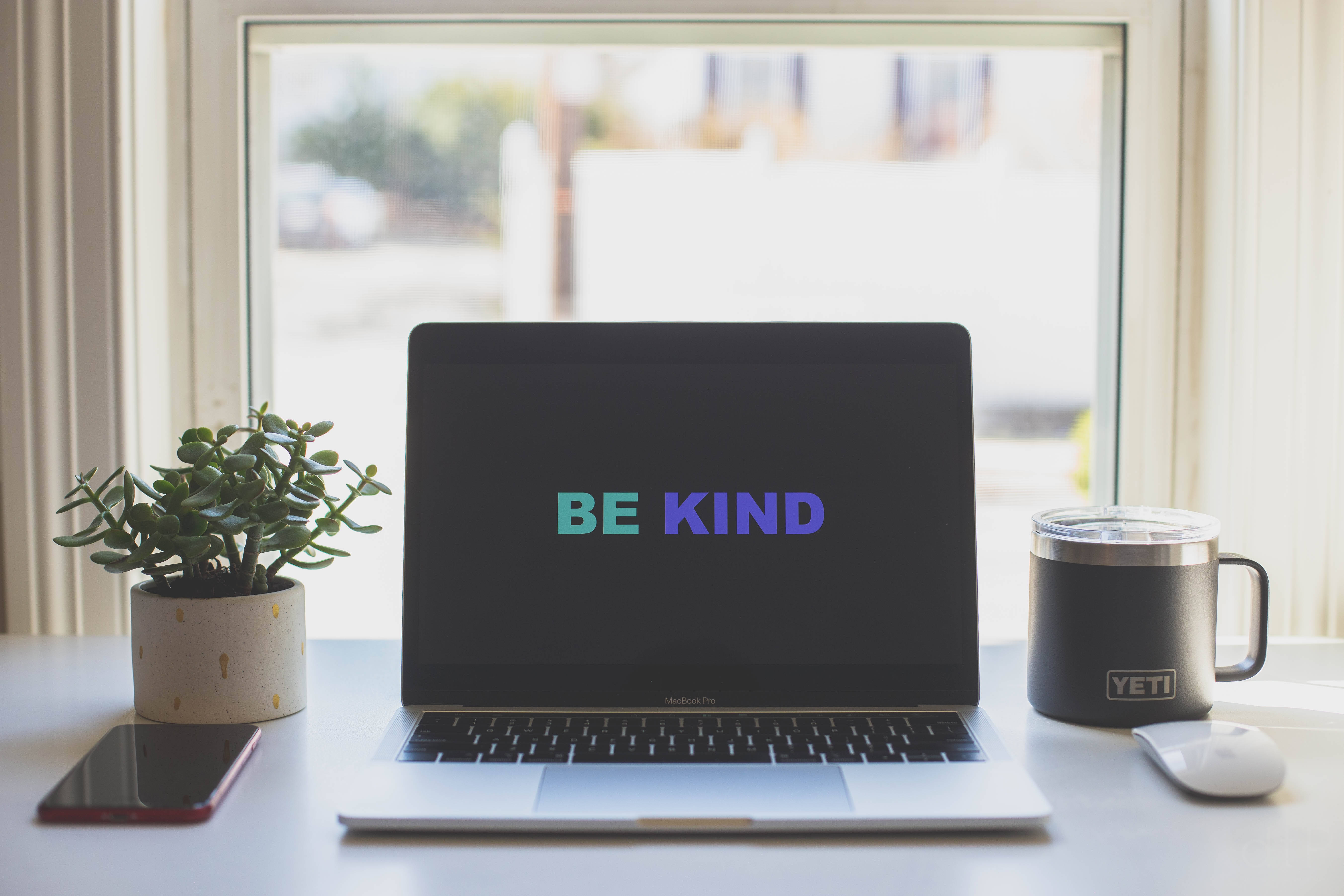 激勵 桌布 壁紙 — 一台顯示「BE KIND」的筆記型電腦放在明亮的窗台上，旁邊有一棵植物、一個Yeti杯子和一部電話。
