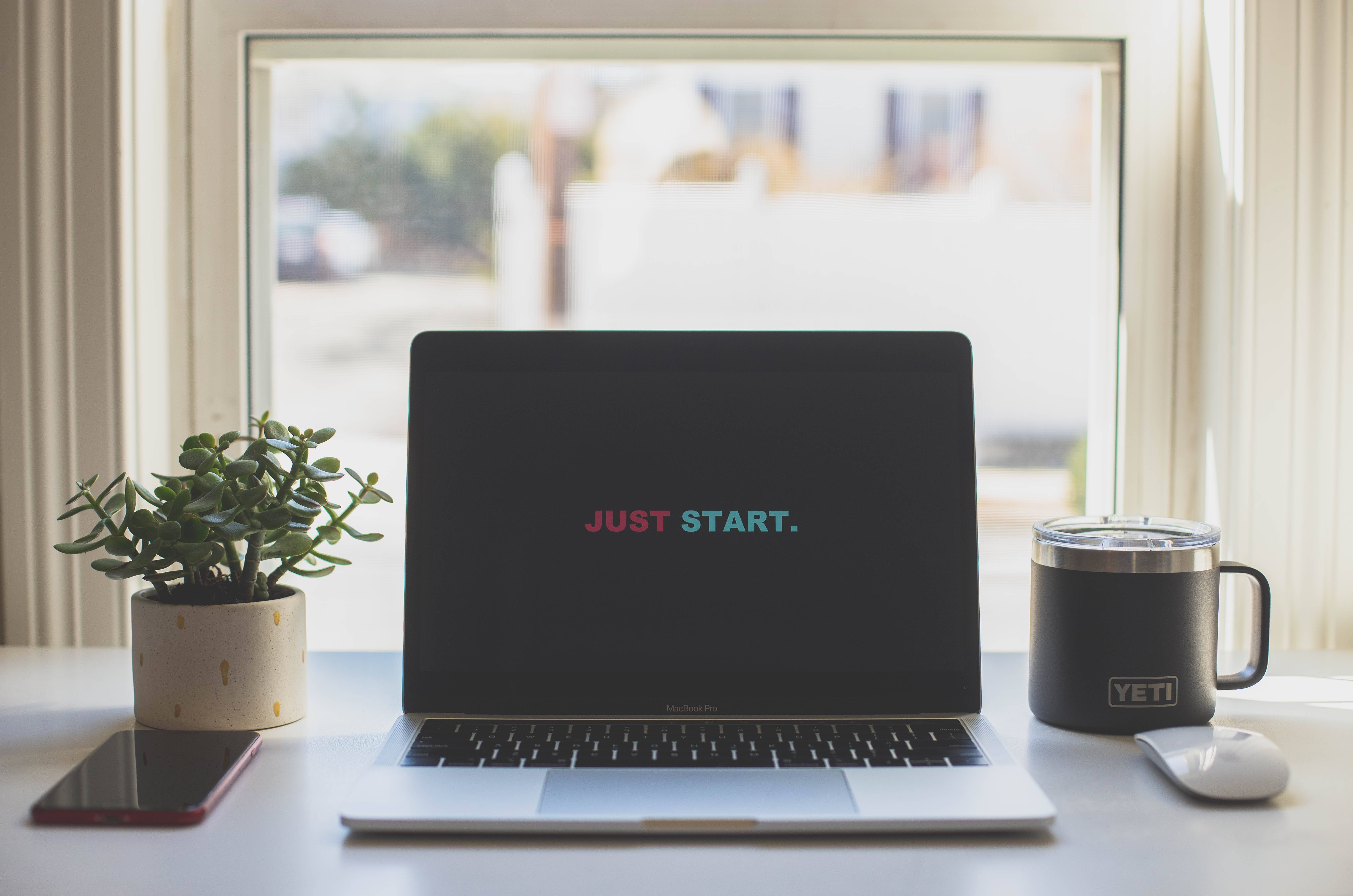 激勵 桌布 壁紙 — 一台顯示「JUST START.」的筆記型電腦放在明亮的窗台上，旁邊有一盆植物、一個Yeti杯子和一部電話。
