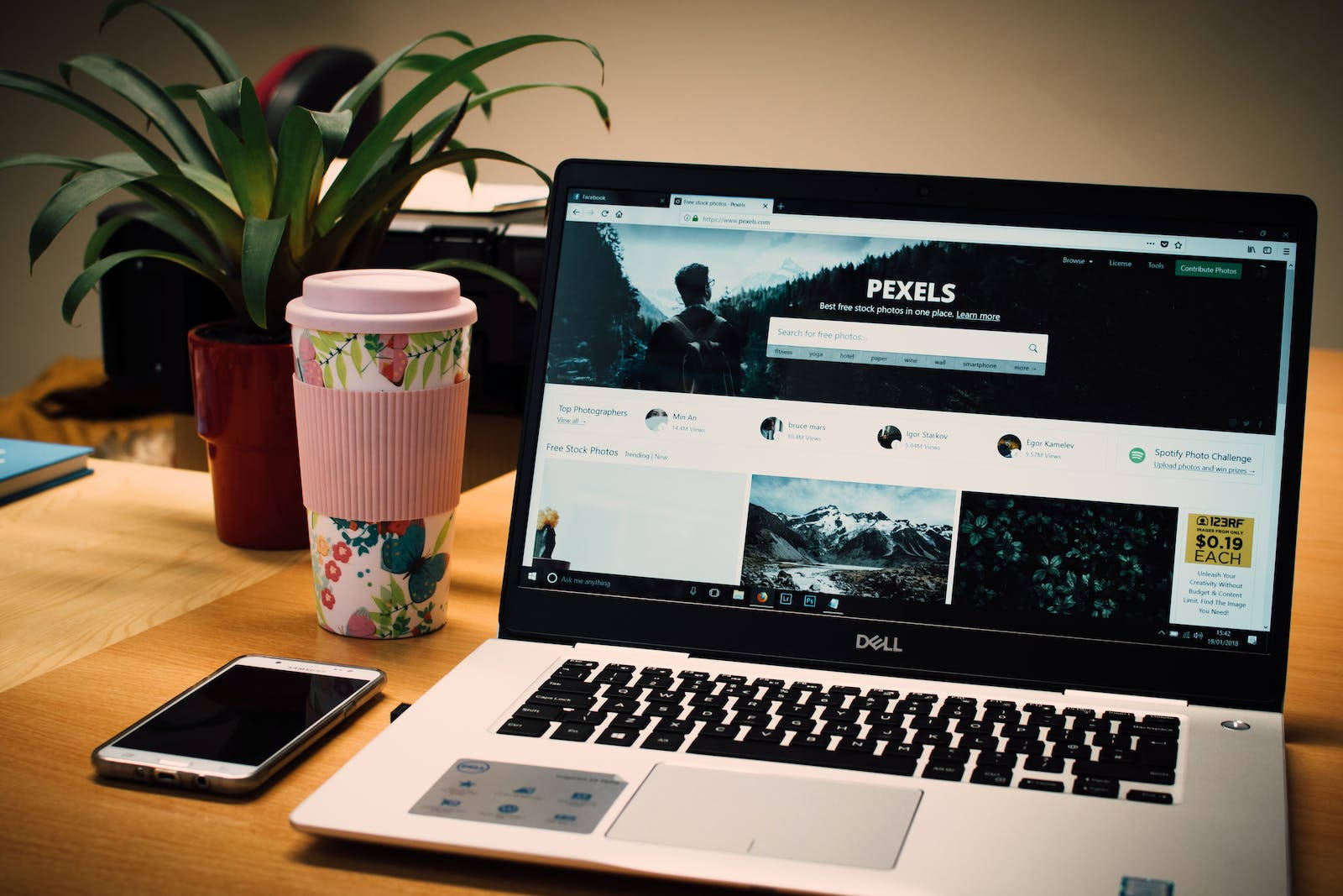 Dell Laptop 桌布 壁紙 — 一台顯示 Pexels 網站的筆記型電腦放在木桌上，旁邊有一個花卉圖案的馬克杯和一盆盆栽。