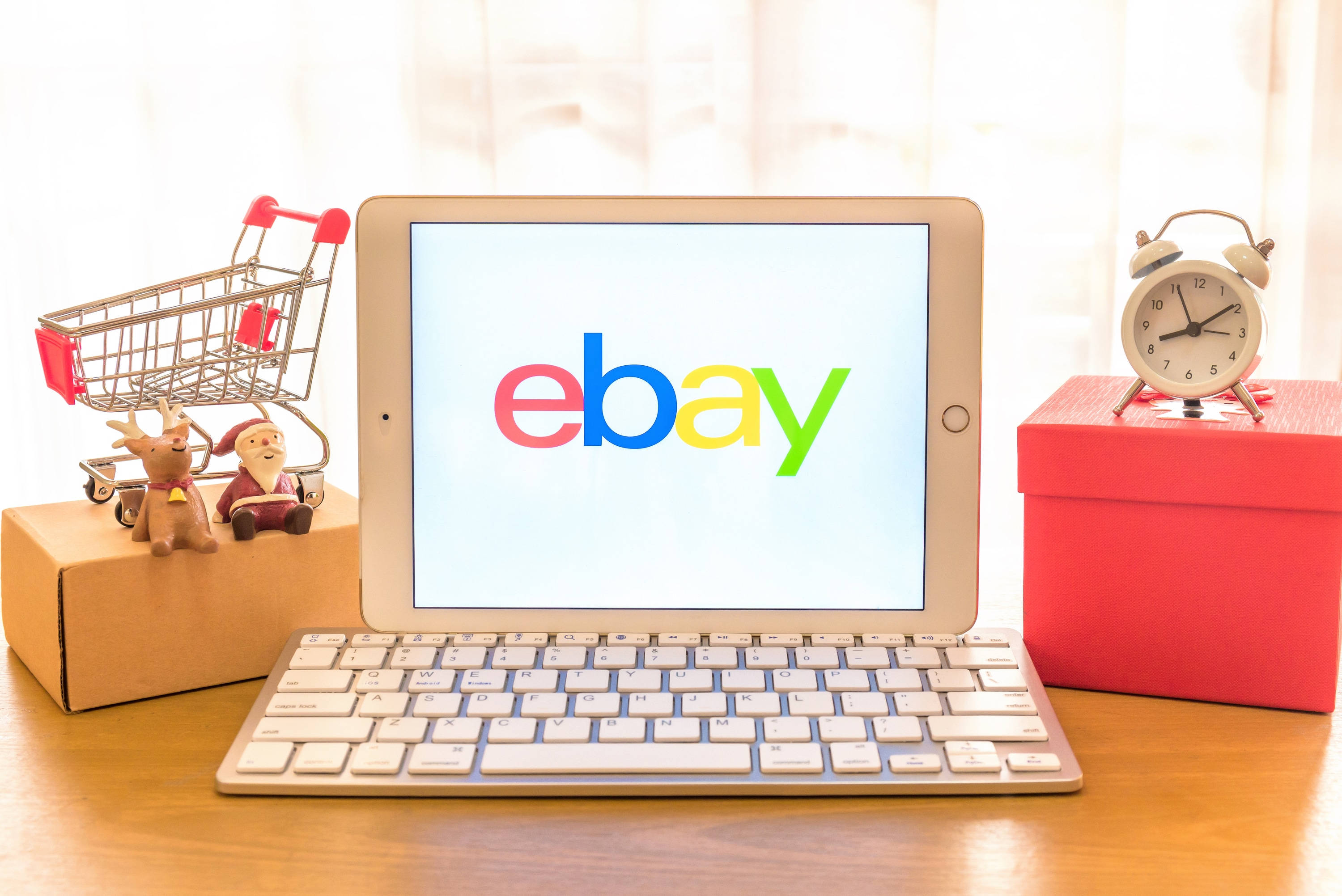 eBay 桌布 壁紙 — 一台顯示eBay標誌的平板電腦放在木桌上，旁邊有一個微型購物車、聖誕老人公仔和一個帶鬧鐘的紅色禮物盒。
