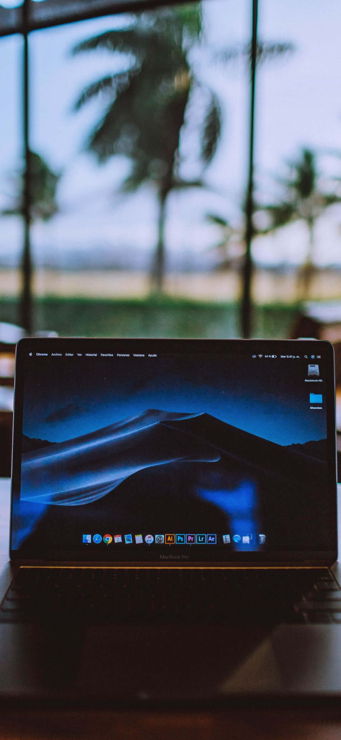 iPhone Xs Computer 桌布 壁紙 — 一台顯示藍色山脈桌面的筆記型電腦放在書桌上，透過後面的窗戶可以看到模糊的棕櫚樹。