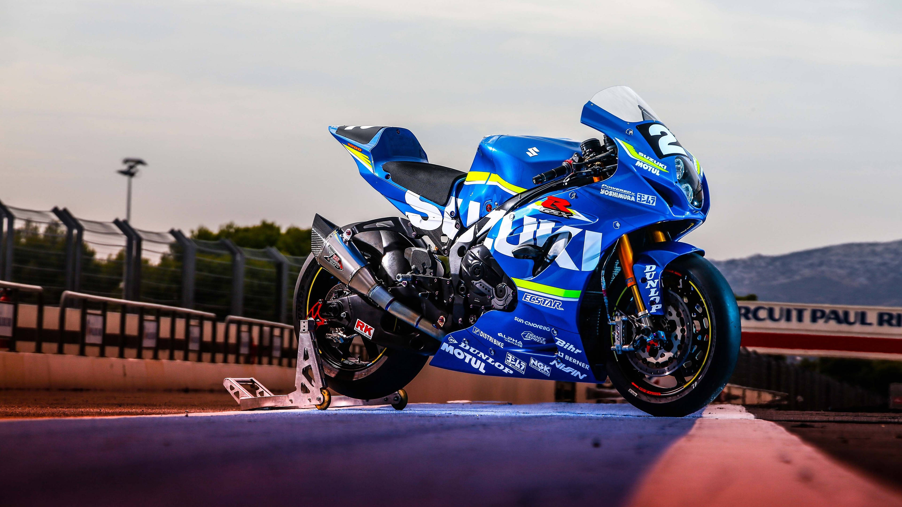 Suzuki 桌布 壁紙 — 一台鮮藍色鈴木賽車機車，車身有2號，在黃昏時分停在賽道上，背景是山脈。