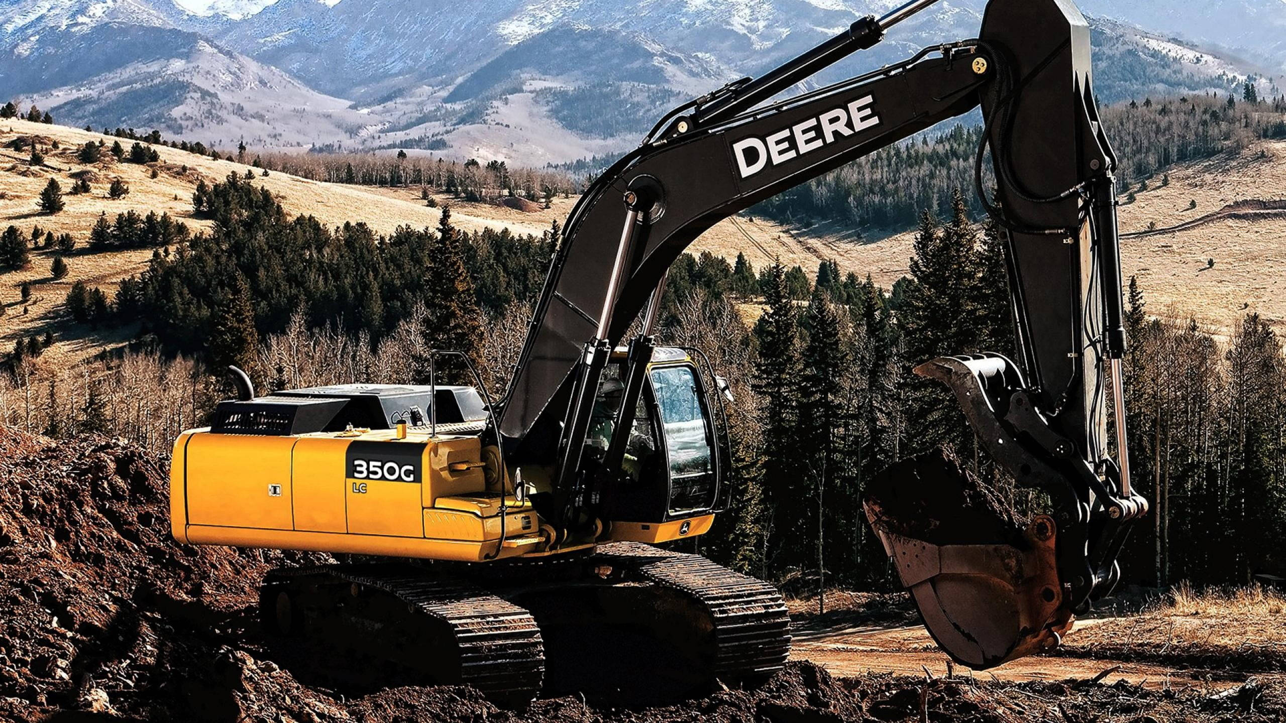 約翰迪爾 桌布 壁紙 — 一台黃色Deere 350G挖土機正在挖土，背景是白雪覆蓋的山脈和常綠森林。