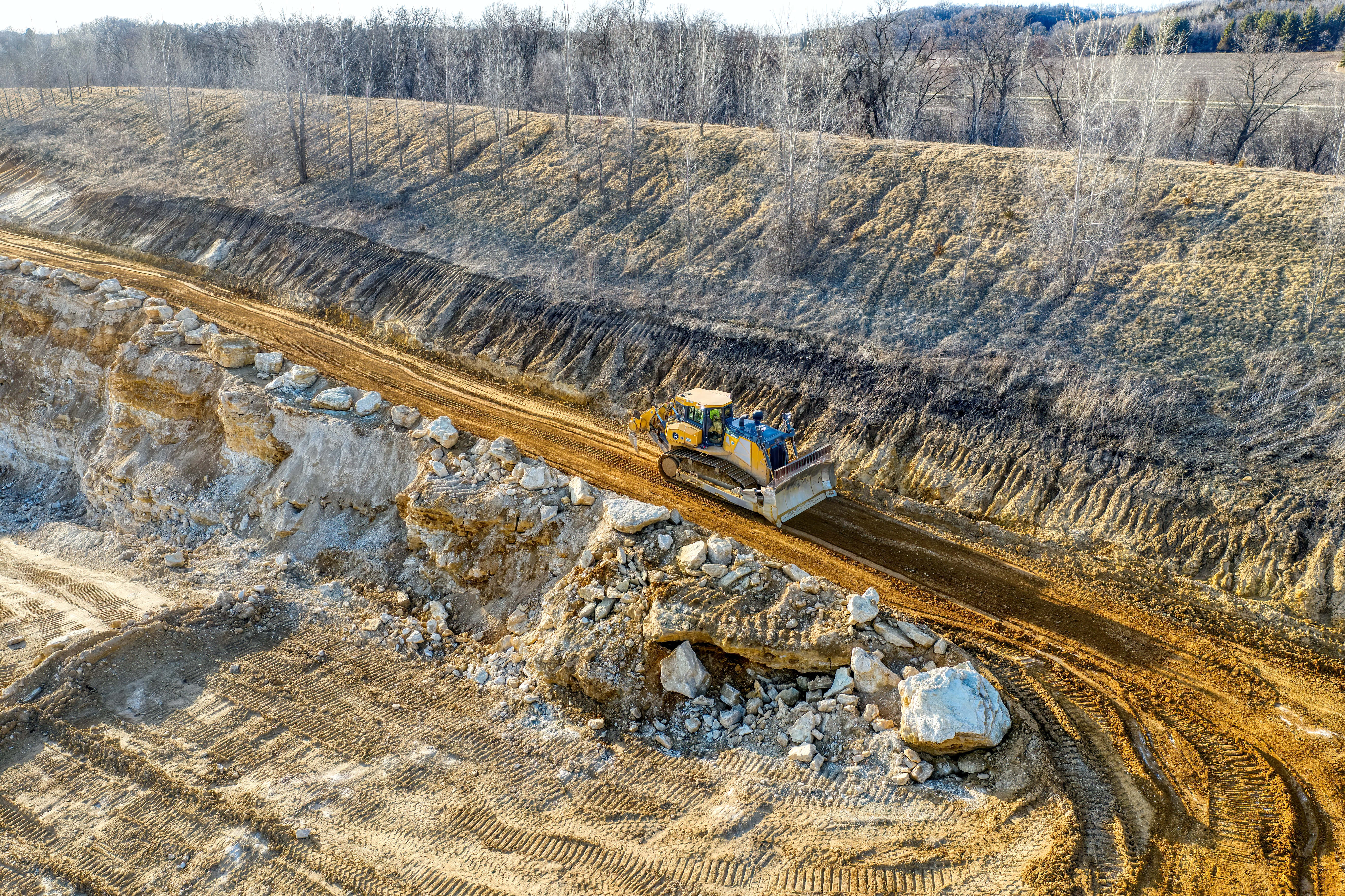 Mining 桌布 壁紙 — 一台黃色推土機在光禿禿的採石場中，沿著崎嶇的泥土路推動泥土，周圍是沒有葉子的樹木，陽光普照。