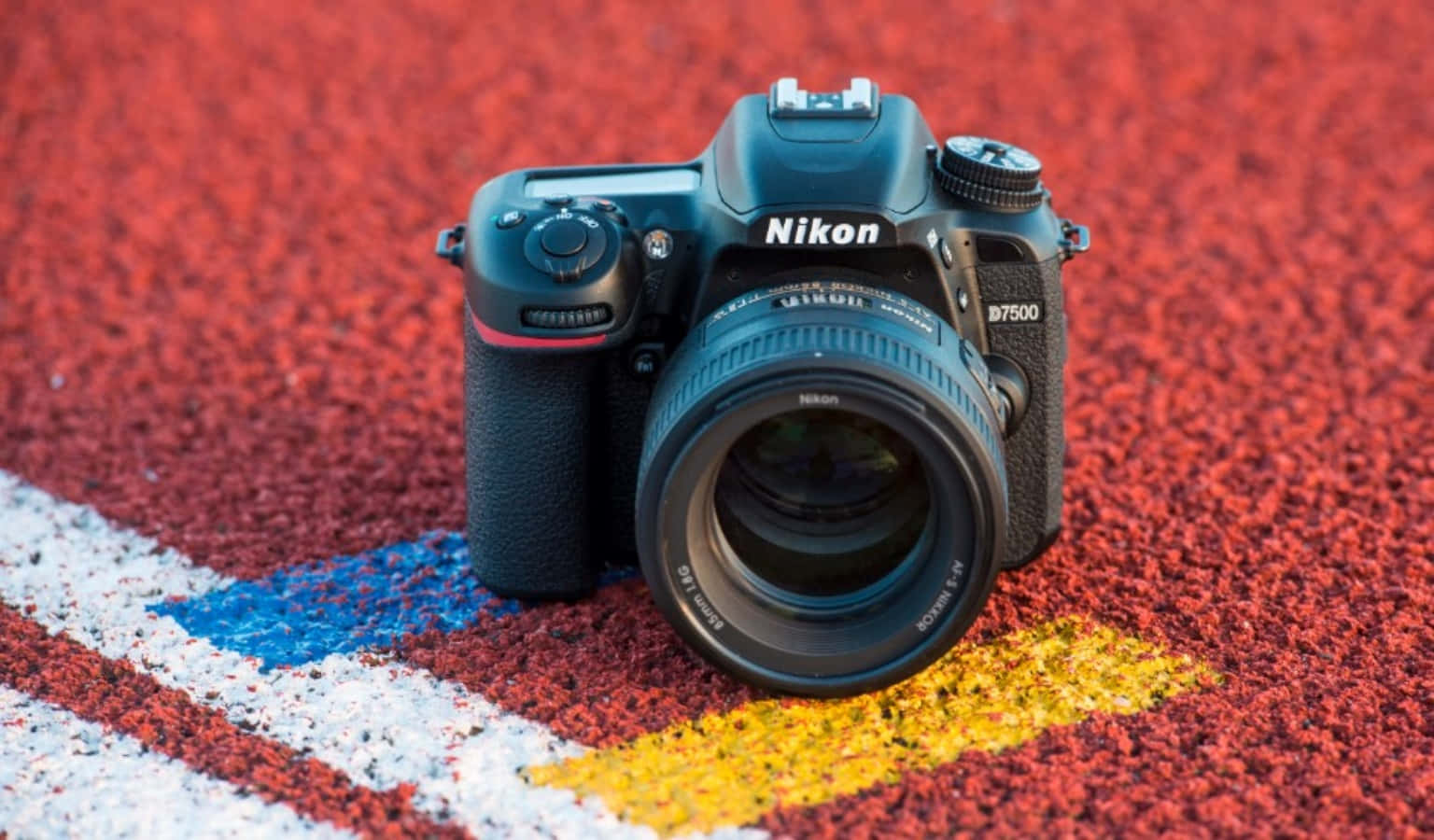 相機 桌布 壁紙 — 一台黑色 Nikon D7500 DSLR 相機放在一條鮮紅色、標有白色、藍色和黃色線條的田徑跑道上。