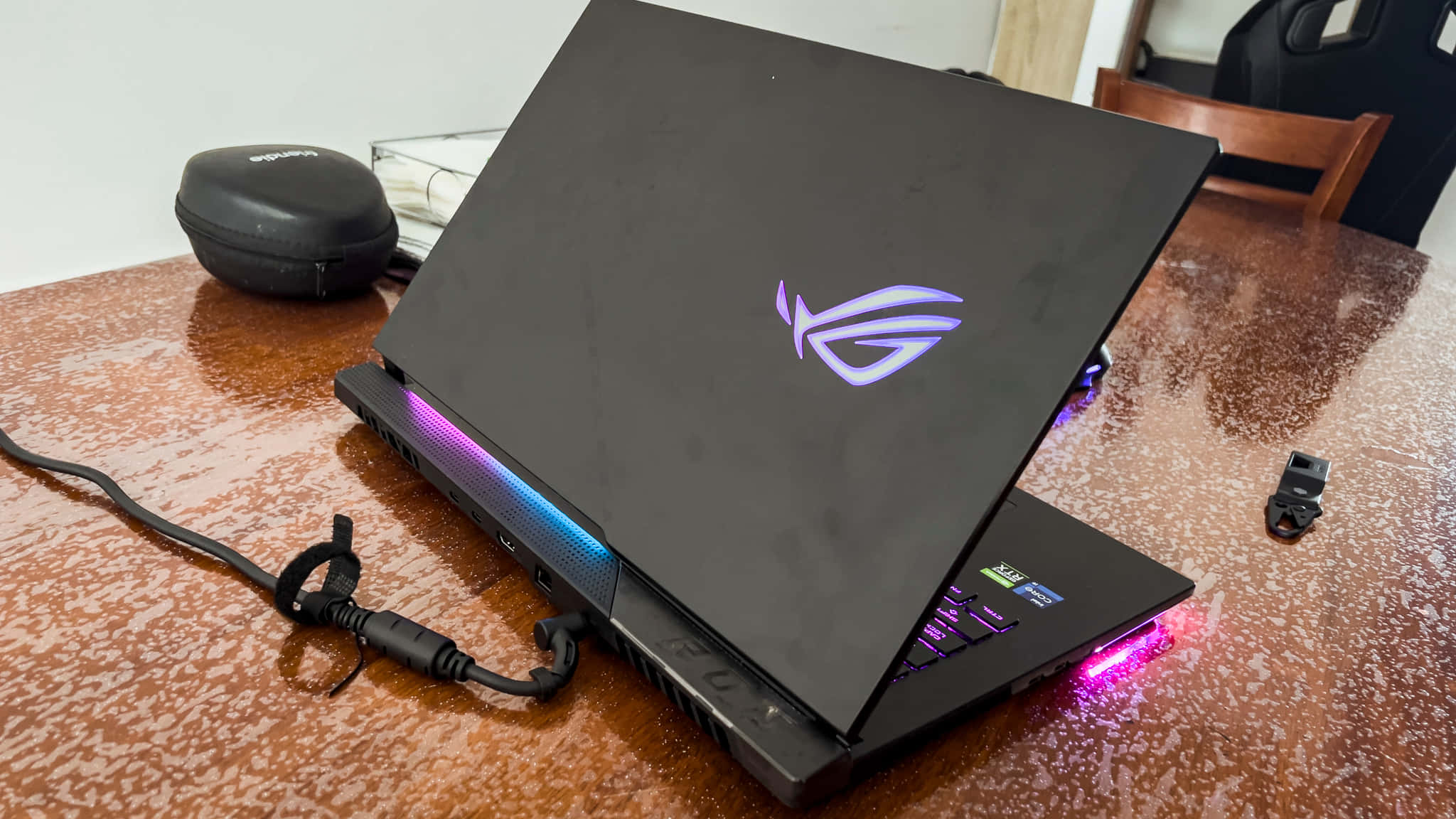 ASUS 桌布 壁紙 — 一台黑色ASUS ROG電競筆電，帶有發光的紫色和藍色RGB燈，打開放在一張有斑點的棕色桌子上，附近有一個耳機和保護套。