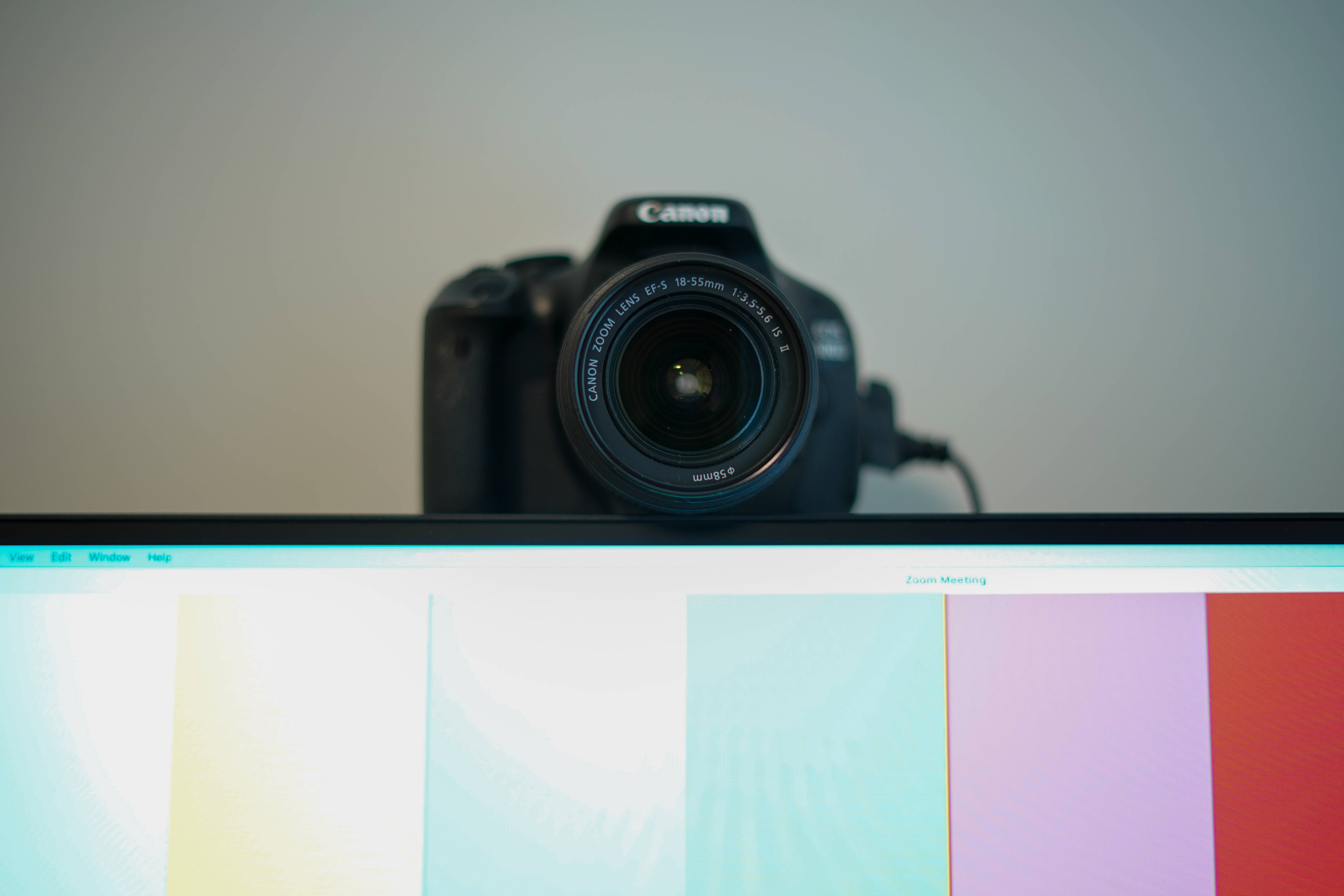 Webcam 桌布 壁紙 — 一台黑色Canon DSLR相機放在顯示器上，顯示器顯示色彩繽紛的Zoom會議背景。