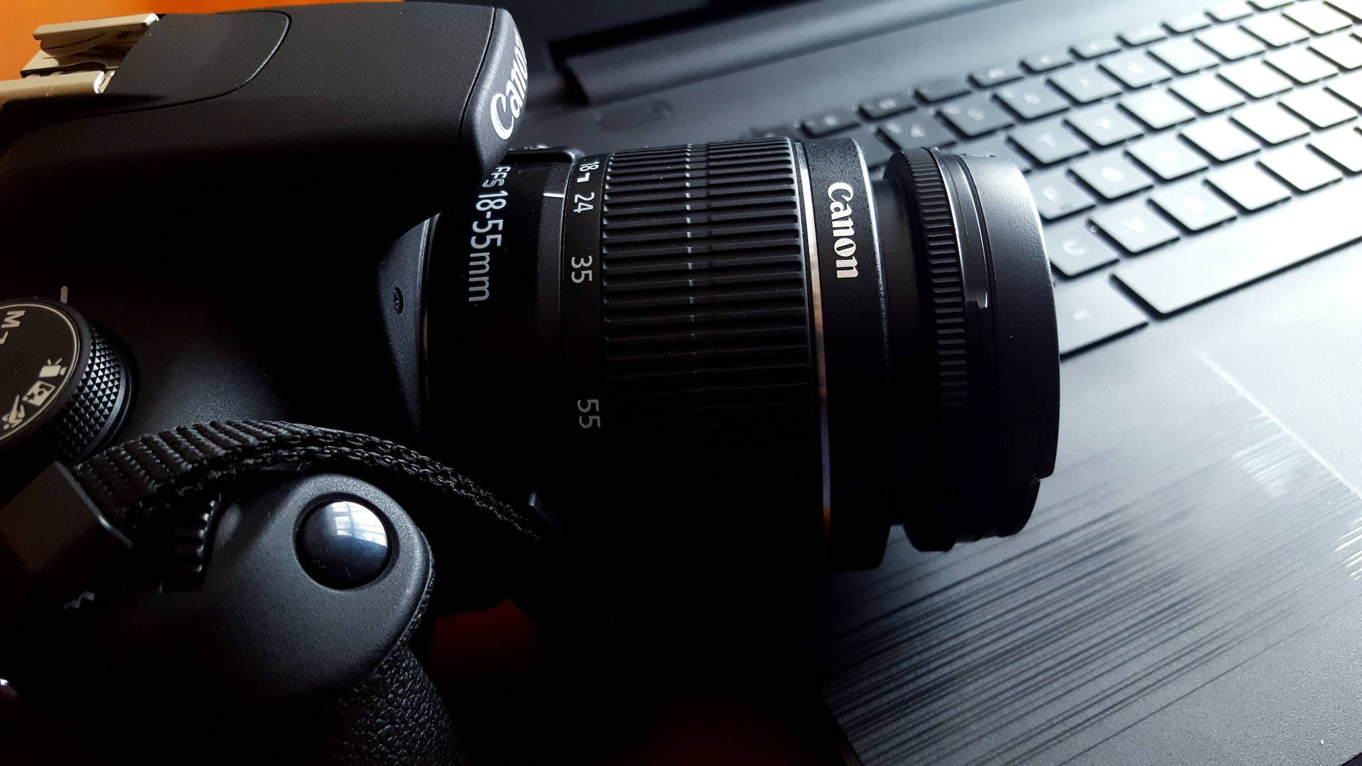 相機 桌布 壁紙 — 一台黑色Canon DSLR相機配備18-55mm鏡頭，放置在昏暗環境中的筆記型電腦鍵盤上。