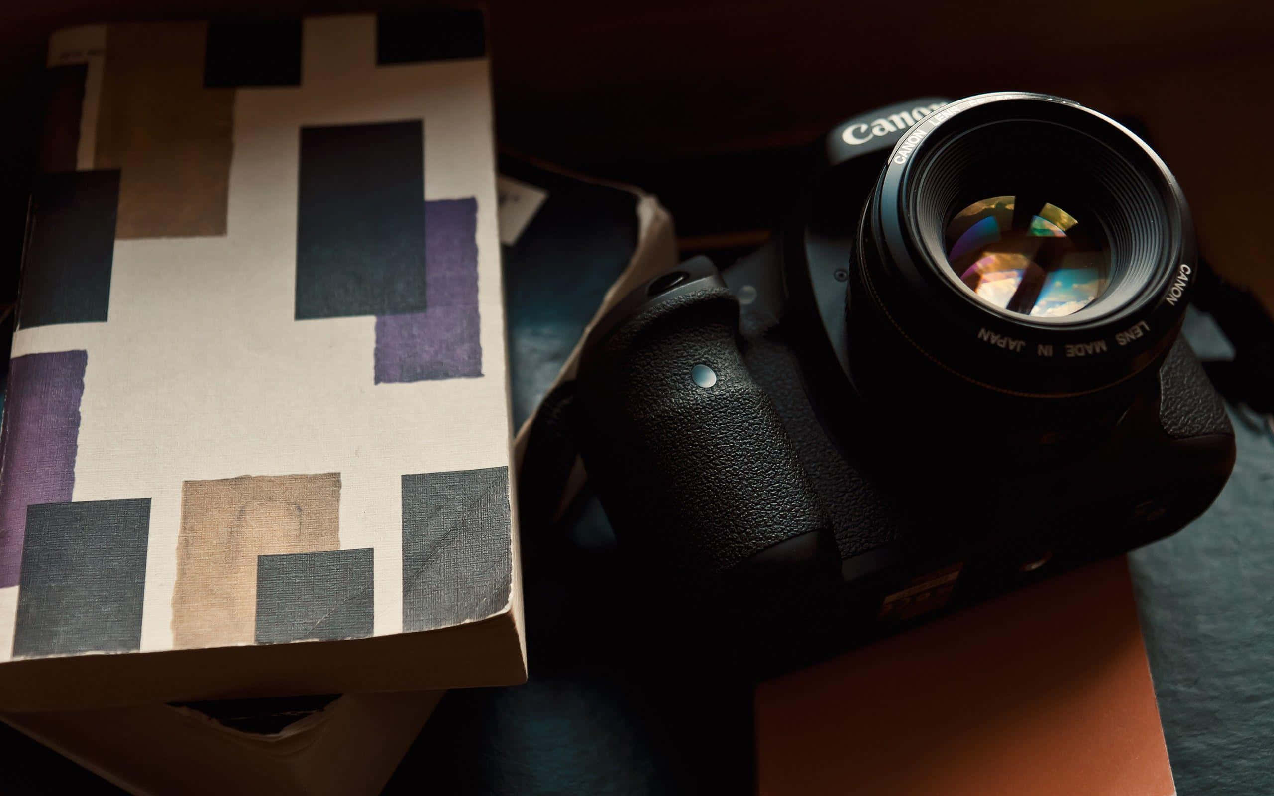 Dslr 桌布 壁紙 — 一台黑色Canon DSLR相機放在一本幾何圖案書旁邊，在溫暖的方向性燈光下。