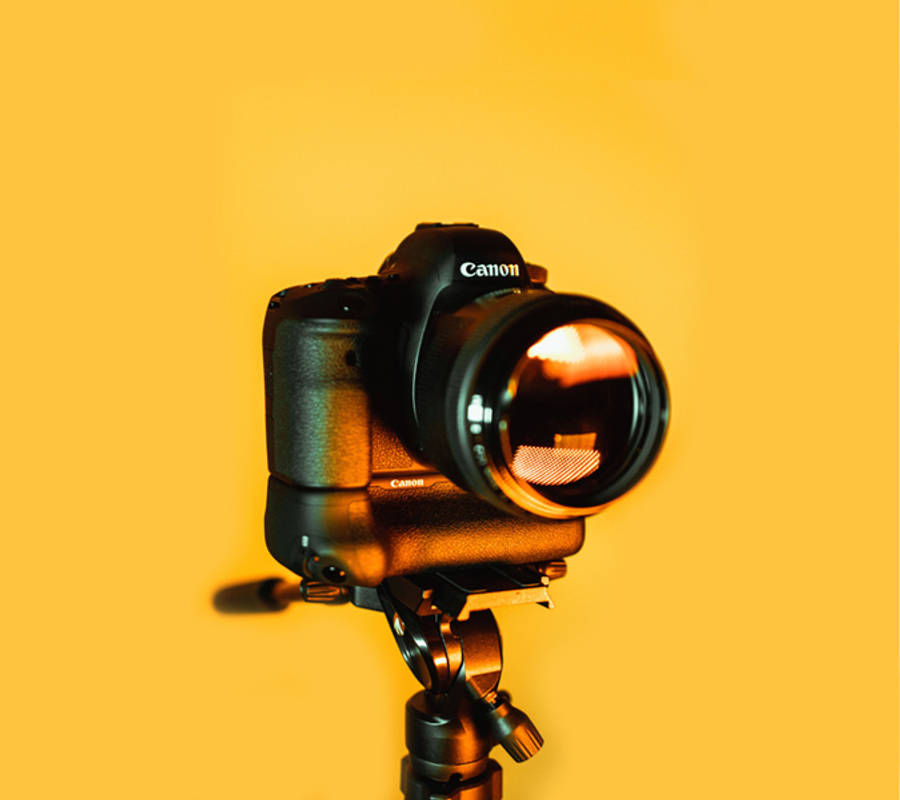 相機 桌布 壁紙 — 一台黑色Canon DSLR相機安裝在三腳架上，背景為溫暖的黃色，光線柔和。