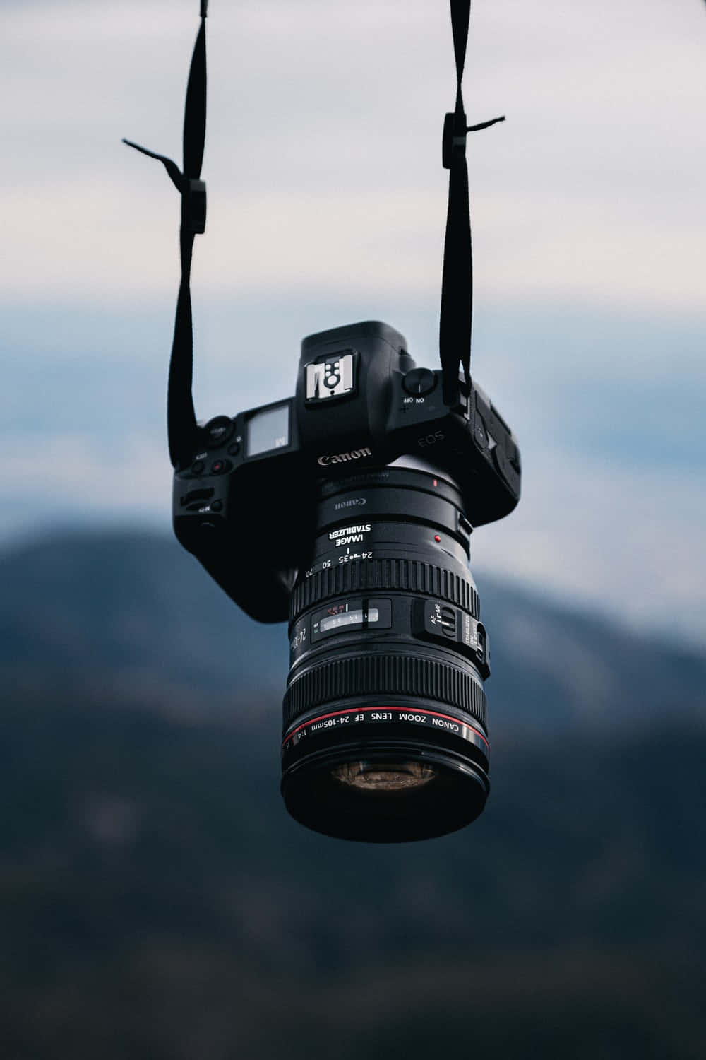 Dslr 桌布 壁紙 — 一台黑色Canon DSLR相機用帶子懸掛在模糊的山巒天空背景下。