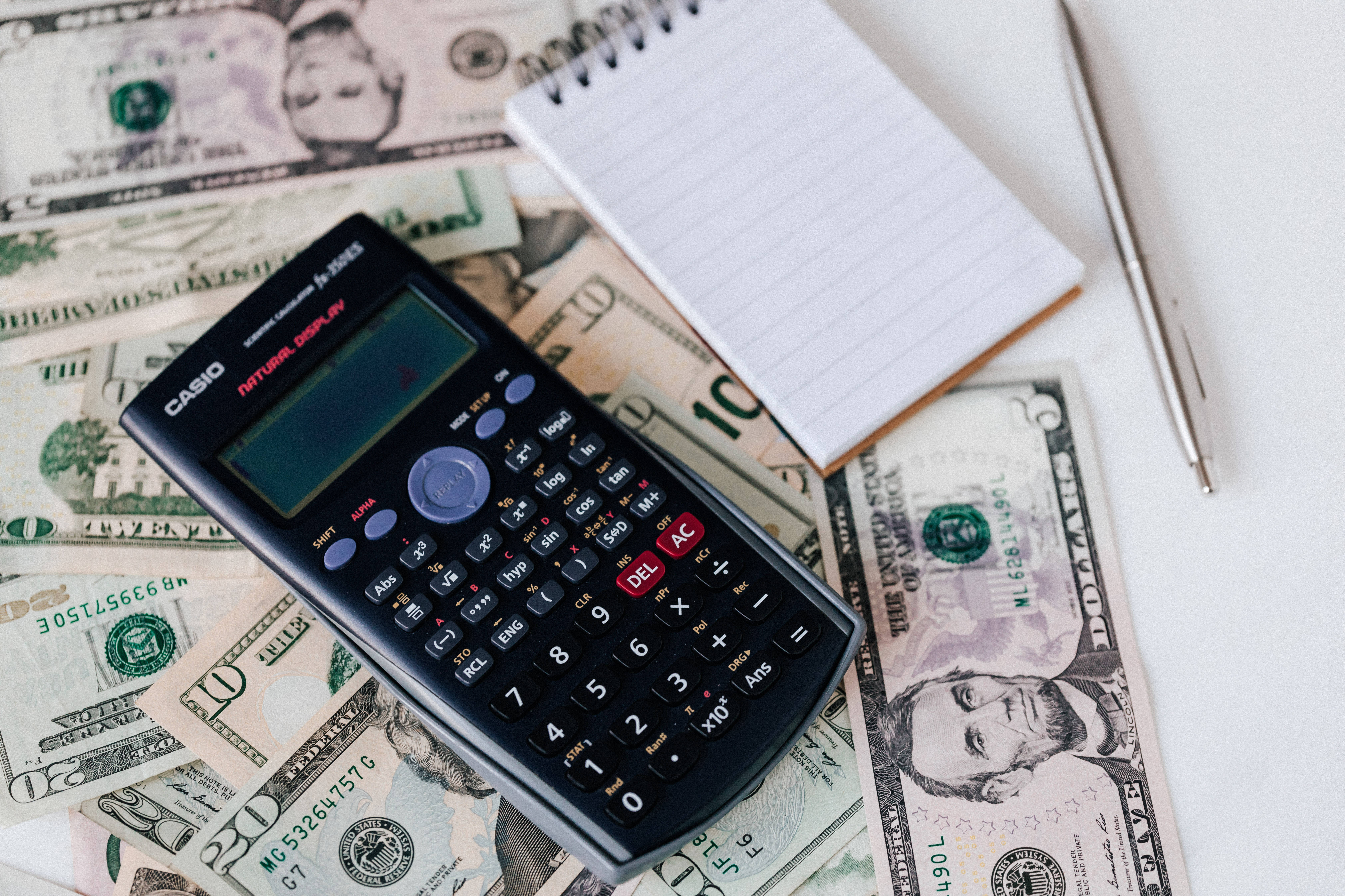 Chartered Accountant 桌布 壁紙 — 一台黑色Casio計算機放在散落的美鈔上，附近有螺旋筆記本和筆。
